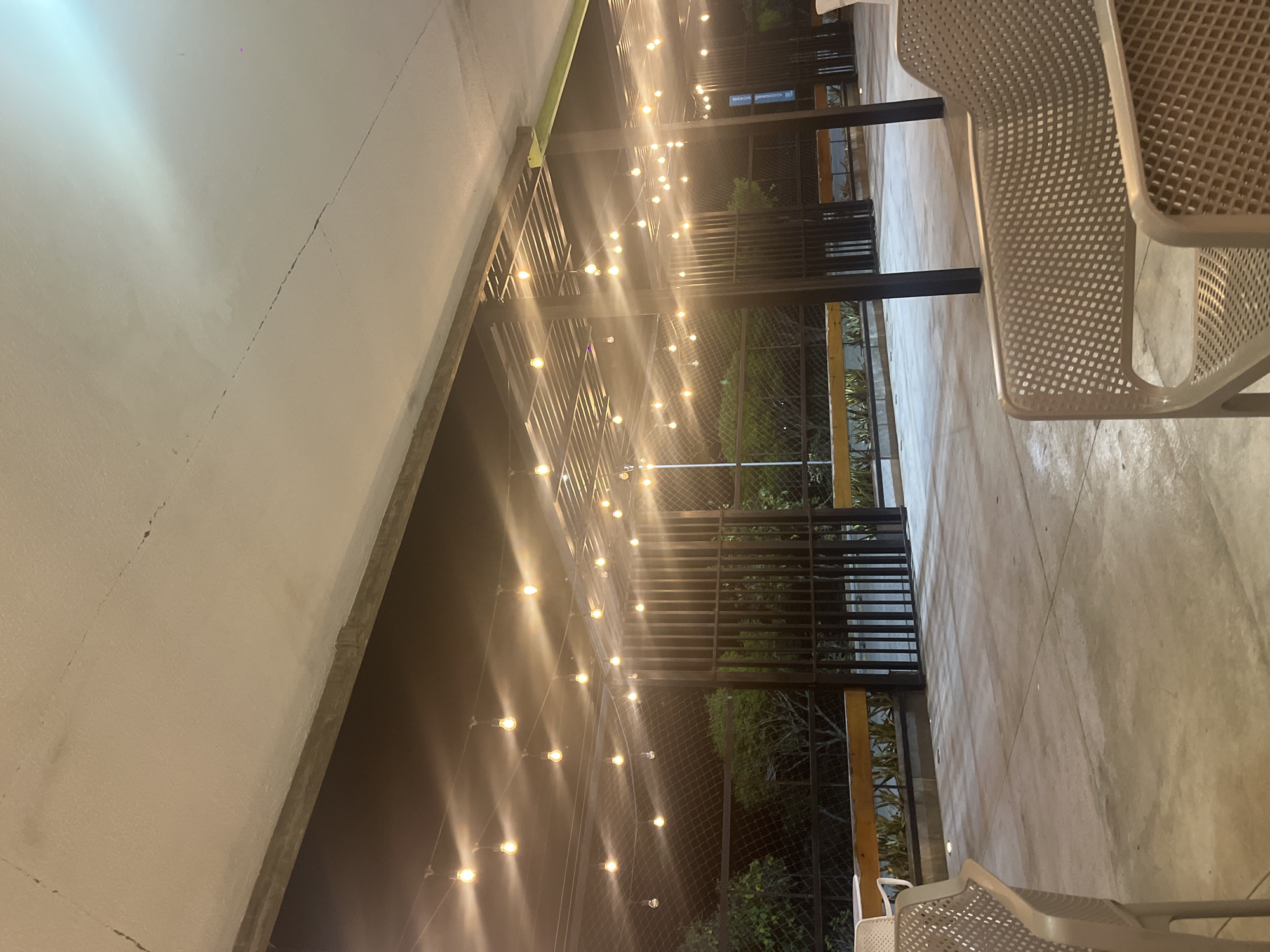 The image captures an outdoor area at night, illuminated by numerous string lights, with the camera positioned looking down a covered walkway or patio. On the left side of the frame, a long, light-colored concrete wall or ceiling runs horizontally, exhibiting a distinct, thin crack along its surface. Overhead, a dark metal grid structure supports multiple strands of string lights, featuring many small, glowing bulbs that cast a warm, yellowish light across the scene. These lights are strung in a crisscross pattern, creating a festive and inviting atmosphere.
To the right, a textured concrete floor or pathway stretches into the distance. In the immediate foreground on the right, two modern, light-colored chairs with a distinctive mesh pattern on their seats and backs are partially visible, suggesting a seating area. Beyond the main metal structure, the background is dark, revealing glimpses of green foliage and a dark mesh fence. Faintly visible in the distance, on a dark sign behind the metal structure, the text "CORRAL KIOS" can be discerned. No people are present, indicating a quiet evening scene, likely a patio or outdoor dining space. The time of day is clearly evening or night due to the prominent use of artificial lighting and the dark surroundings.