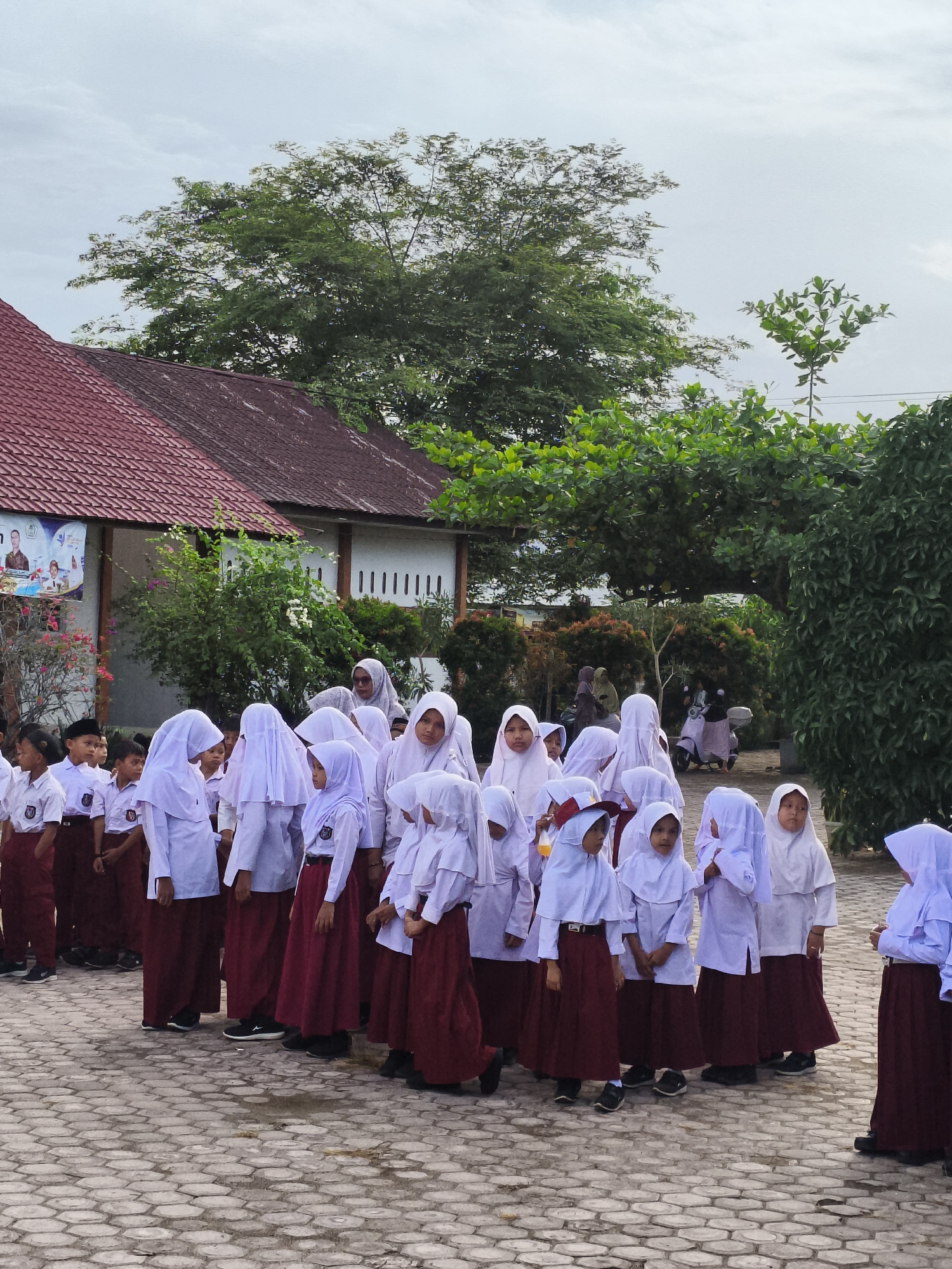 In Baratpayaitek, Indonesia, a group of schoolchildren, predominantly girls, stand in an outdoor courtyard. They are dressed in school uniforms consisting of white shirts, white hijabs, and maroon skirts. The children are arranged in multiple rows, with some looking towards the camera and others looking away. The background features a building with a red roof, trees, and a cloudy sky. A poster is visible on the wall of the building. The ground is paved with hexagonal stones. A few adults are also present in the background, and a scooter is parked nearby. The overall impression is a gathering of students at their school.