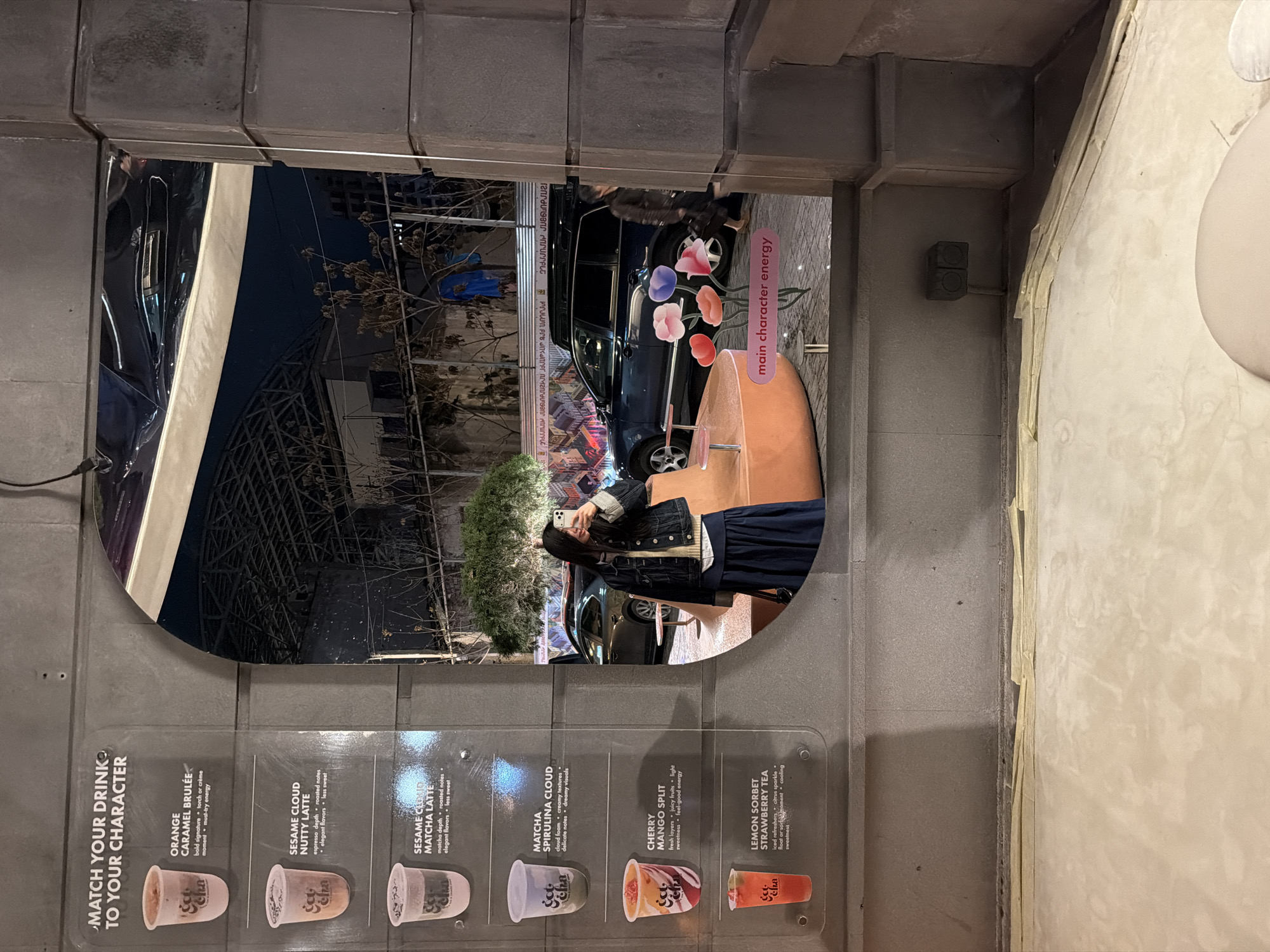 The image captures a scene from inside a building looking out through a large, irregularly shaped window at night in Yerevan, Armenia. The foreground features a textured, grey concrete wall. On the left side of the wall is a clear acrylic menu display titled "MATCH YOUR DRINK TO YOUR CHARACTER." It lists six different beverages: "ORANGE CARAMEL BRULÉE," "SESAME CLOUD NUTTY LATTE," "SESAME CLOUD MATCHA LATTE," "MATCHA SPIRULINA CLOUD," "CHERRY MANGO SPLIT," and "LEMON SORBET STRAWBERRY TEA." Each drink is accompanied by a description and an image of a cup with the "See Cha" brand logo.
Through the window, a dark urban environment is visible. A person, appearing to be a young woman with long dark hair, wearing a denim jacket and a dark skirt, is standing outside, holding a smartphone up to take a selfie. Behind her is a dark-colored SUV car. To the right of the car, a pink sign with colorful floral cutouts reads "main character energy." Further in the background, a banner with text in an unfamiliar script is visible, along with architectural structures, possibly parts of a building or a bridge, and some sparse vegetation. The overall impression is an evening or nighttime setting, indicated by the darkness outside and artificial lighting within the scene.