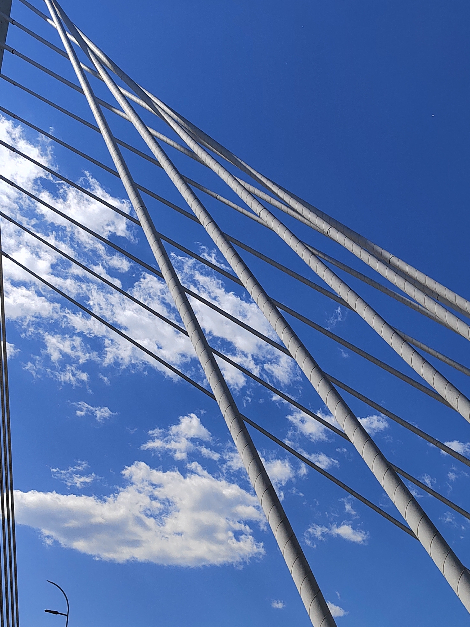 The image shows a low-angle view of the Millennium Bridge in Podgorica, Montenegro. Several white cables are visible, spanning diagonally across the frame against a bright blue sky interspersed with white, puffy clouds. The cables are of two types: thicker, vertically oriented cables with a cylindrical appearance, and thinner, horizontal cables. A portion of the bridge's structure is visible in the lower-left corner, along with a street light in the bottom center of the frame.