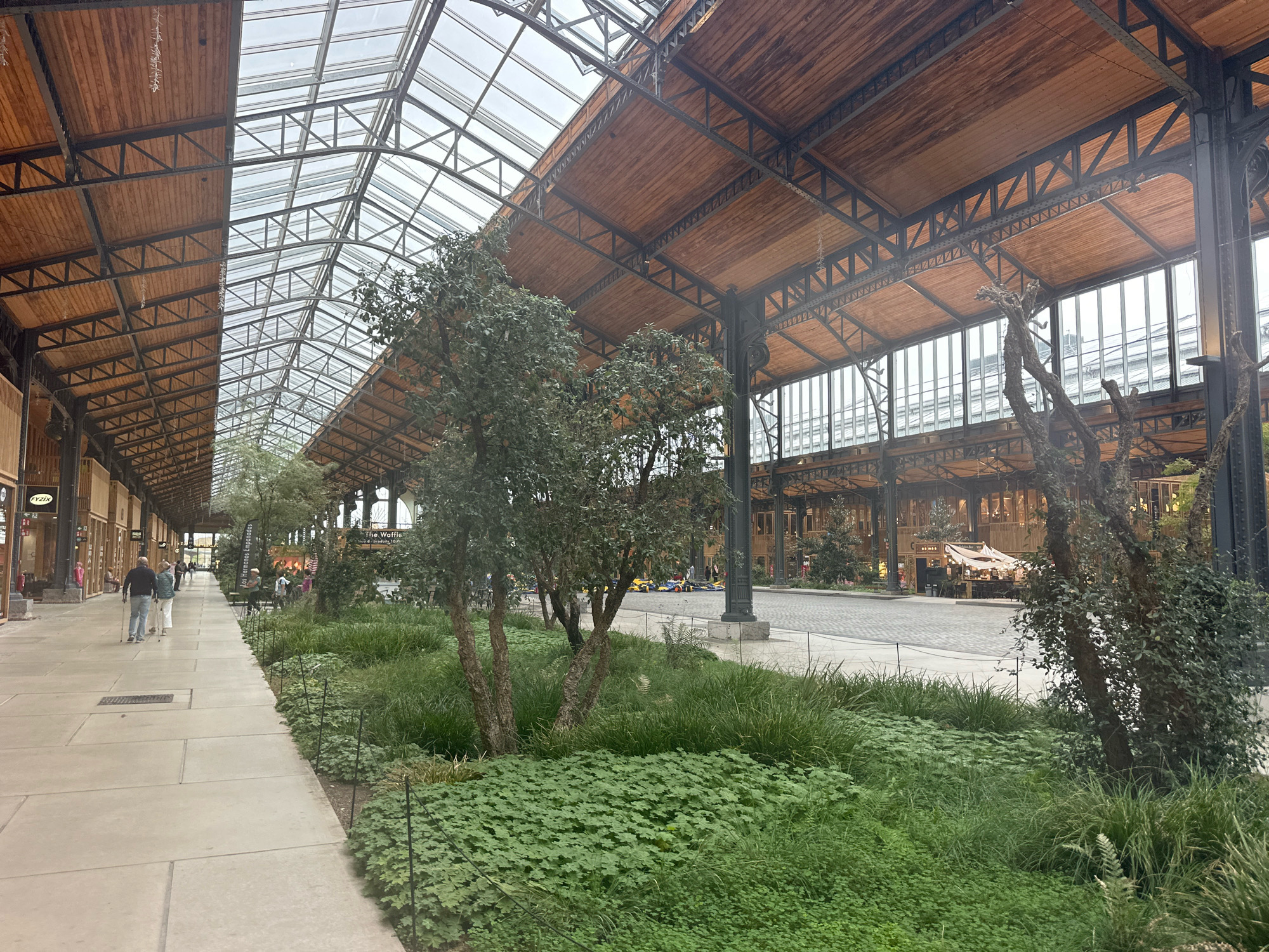 The image shows an interior view of the Tour & Taxis building in Brussels, Belgium, a former customs house that has been redeveloped into a multi-purpose venue. The architecture features a vast glass roof supported by a metal and wooden framework, allowing natural light to flood the space. On the left, a paved walkway runs alongside a row of shops and businesses, indicated by signage such as "LYZIX." Several people are walking along the pathway, including a couple with walking sticks. To the right of the walkway, an area is landscaped with lush green vegetation, including trees, tall grasses, and ground cover. In the background, the central area of the building appears to be an open space with stalls or small shops set up. Further in the distance, more of the building's interior structure and large windows are visible. The overall impression is of a bright, airy, and modern commercial and public space within a historic industrial building.