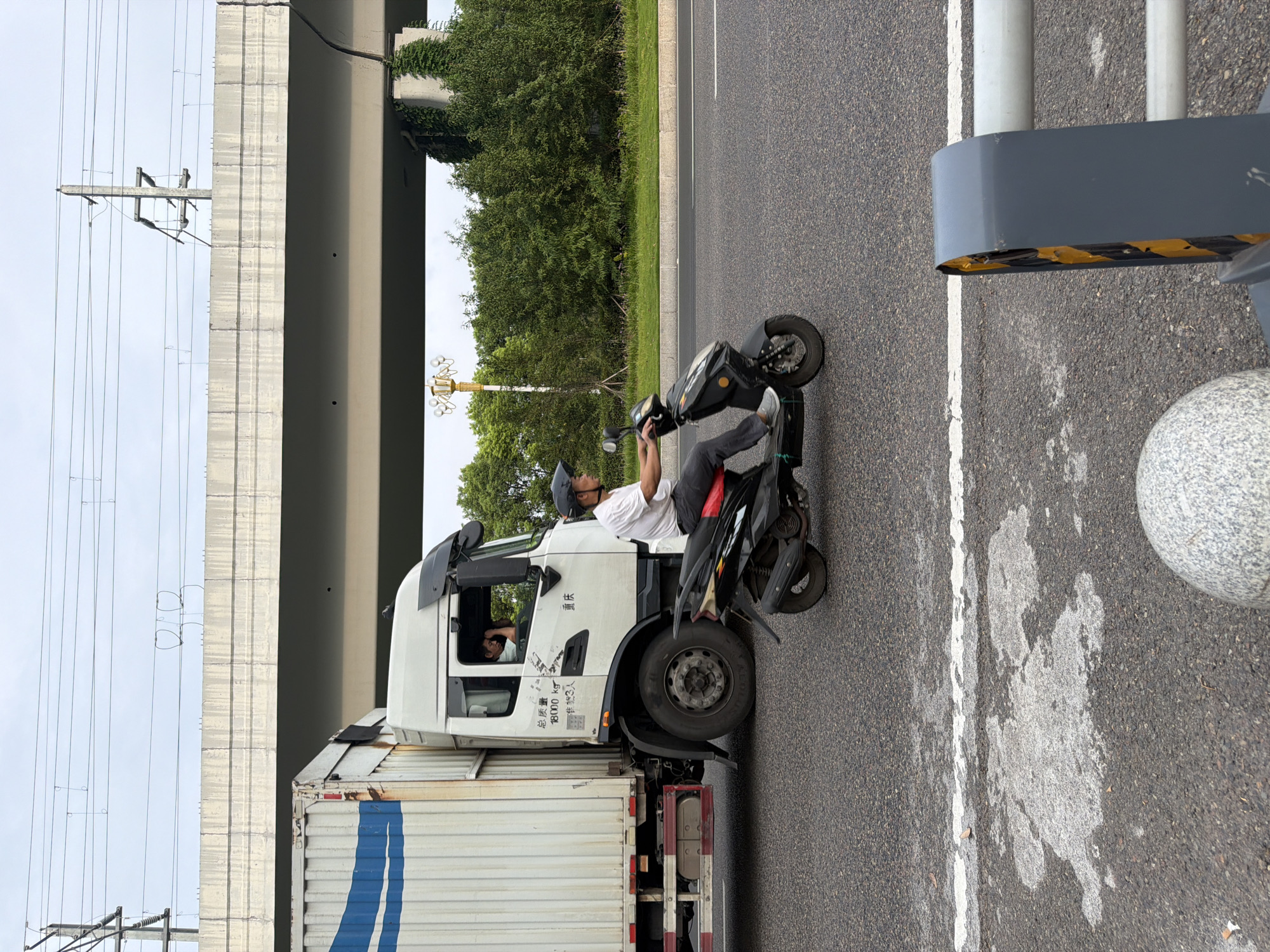 In Jiangjin, China, a white truck with a blue stripe on its cargo container is traveling on a road. The truck has Chinese characters and "18000 kg" printed on its side. A man wearing a helmet and a white t-shirt is riding a black scooter next to the truck. He is looking towards the truck. In the background, there is an elevated concrete structure, possibly an overpass, with power lines running above it. Lush green trees and foliage are visible on the side of the road. The sky is overcast.