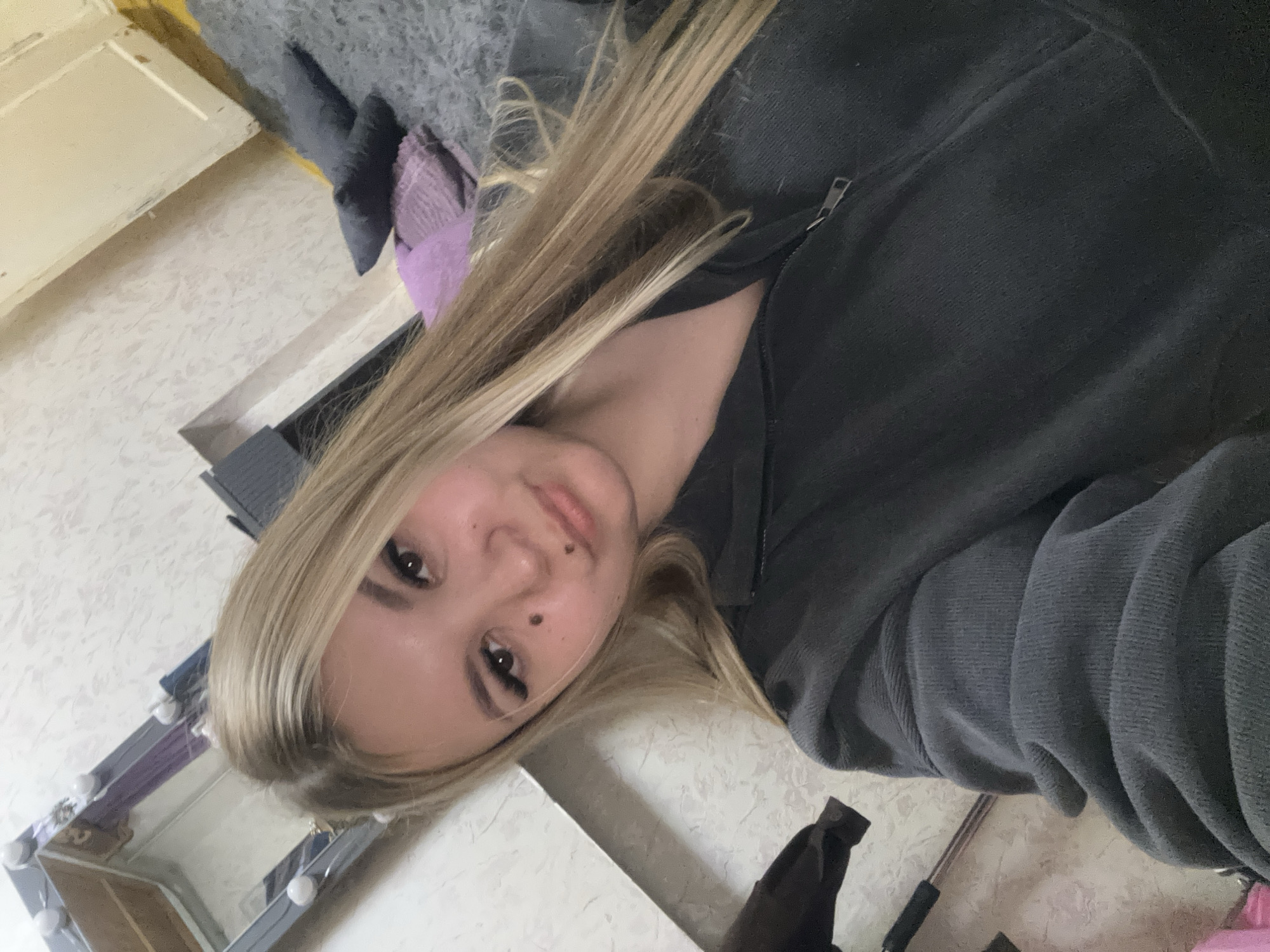 The image shows a young woman with blonde hair, taking a selfie indoors. She is wearing a dark grey zippered hoodie. Her face is angled slightly to the right, and her eyes are looking towards the camera. She has a mole on her left cheek and another on her right cheek, near her nose. The background appears to be a bedroom, with a mirror with lights visible in the upper left corner. A bed with dark grey and purple pillows is in the background to the right. The overall lighting suggests an indoor setting, possibly during the daytime. There is no indication that this image is located in the city of Большой Букор, Russia.