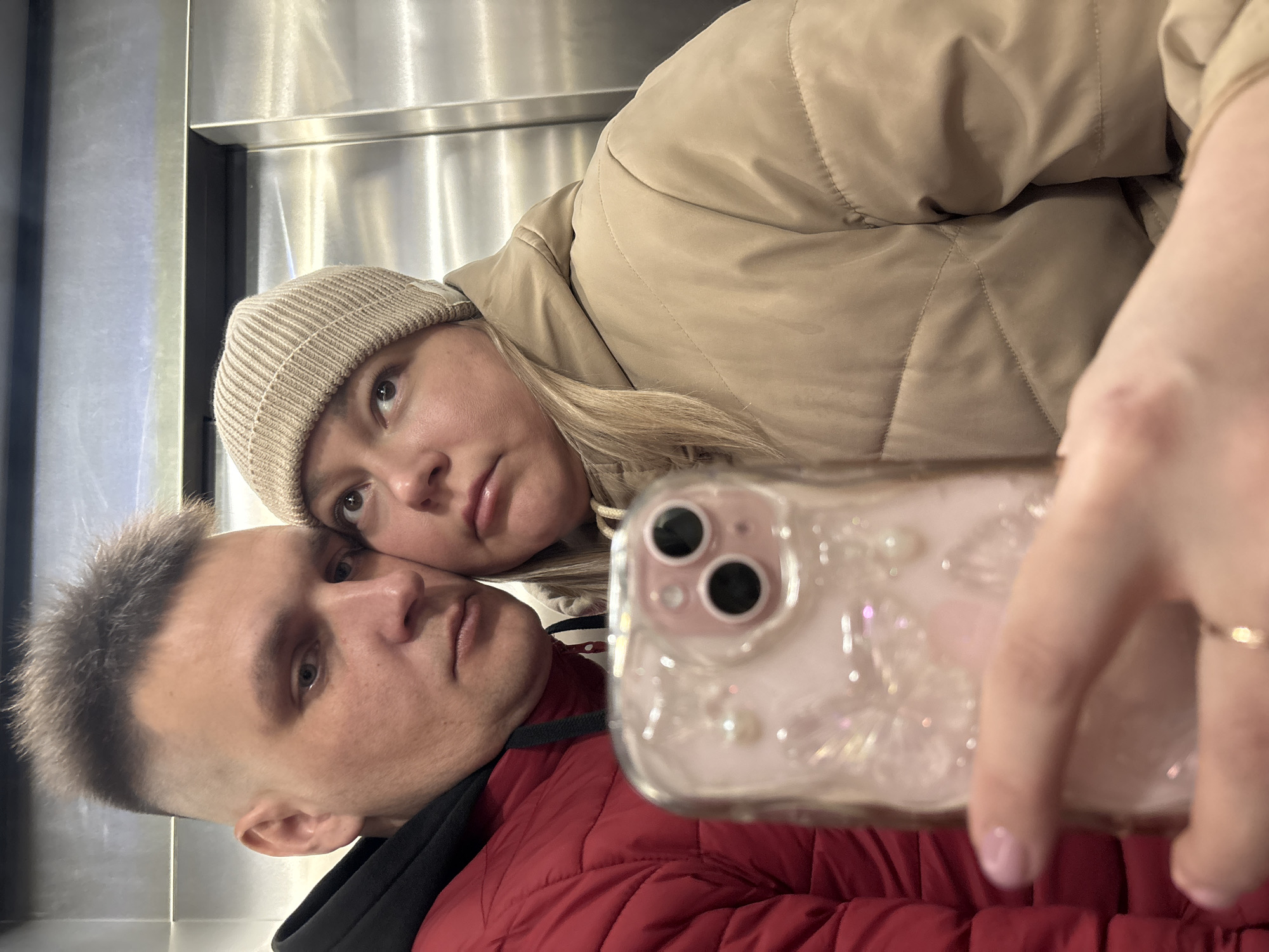 A couple is taking a selfie in what appears to be an elevator. The man, wearing a red puffer jacket, is positioned below the woman. The woman, wearing a beige puffer jacket and a light-colored beanie, leans her head towards the man. They are holding a smartphone in front of them to capture the image. The elevator has stainless steel walls. The location is identified as Izhevsk, Russia.