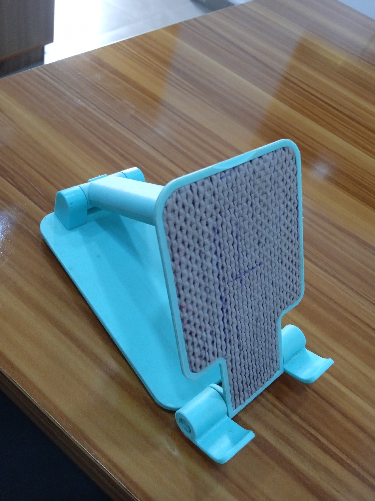 A close-up shot shows a light blue plastic phone stand on a brown wooden table. The stand features a textured gray backing with dark vertical lines, possibly markings or a design element. The plastic components have a glossy finish, reflecting light. The table's surface has a distinct wood grain pattern.