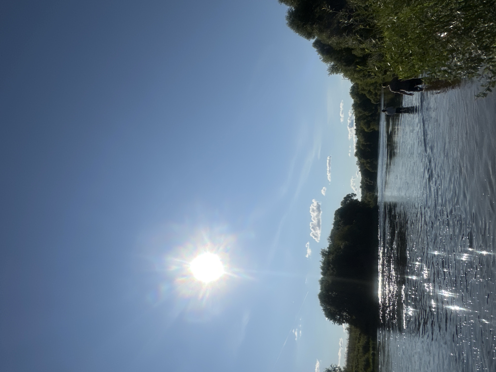 In Kozino, Russia, on a clear day with a bright sun, two individuals are walking through a wide, calm river. The sun casts a strong glare on the water's surface, creating sparkling reflections. The riverbanks are lined with lush green trees and vegetation, with a small island or peninsula covered in dense trees visible in the mid-ground. The sky is a vibrant blue with a few scattered white clouds. The individuals are wading through the shallow water, their figures silhouetted against the bright reflections.