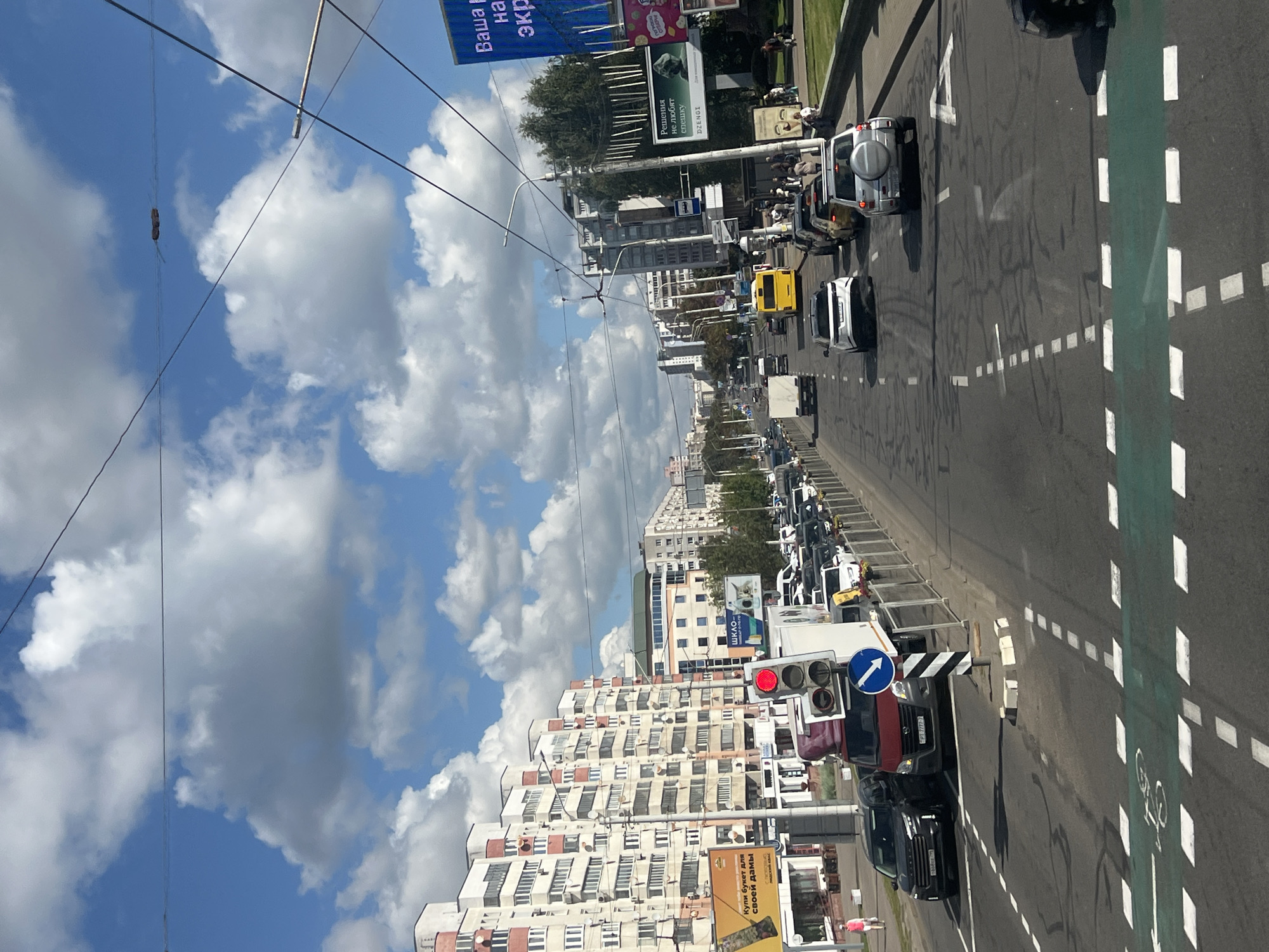 This is an eye-level, forward-facing, exterior, daytime, medium shot photograph taken from a vehicle traveling on a multi-lane road in Minsk, Belarus. The scene depicts an urban street with moderate traffic.
In the foreground, the road surface is visible with painted white dashed lines indicating lanes and a green-painted bicycle lane crossing. Several cars are driving or stopped in traffic. A traffic light is visible on the right side of the road, showing a red light.
The middle ground shows a street lined with tall, multi-story residential buildings on the left, characterized by rows of windows and balconies. On the right side, there are more buildings, some with commercial signage, trees, and a sidewalk where pedestrians are visible. A yellow bus and several other vehicles are on the road.
The background shows a cityscape with more buildings and a bright blue sky with scattered white clouds. Overhead power lines are present, suggesting the presence of trams or trolleybuses. Billboards with advertisements are also visible on the right side.
The atmosphere appears to be a typical urban day with clear weather. The focus is on the street scene and the surrounding architecture.