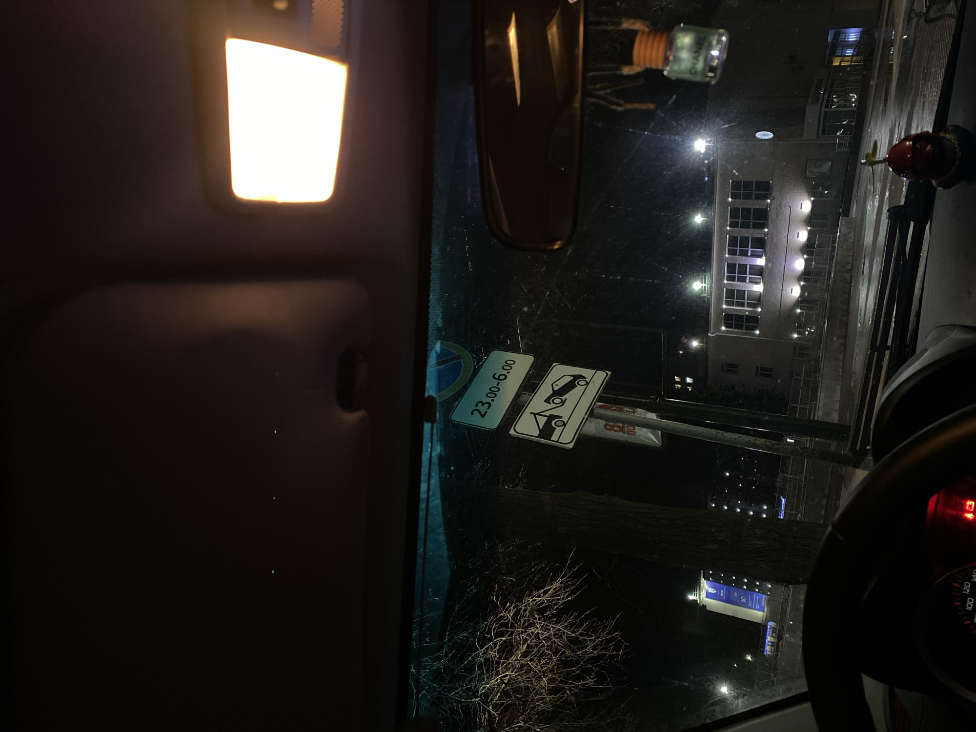 Captured from inside a car at night, the image shows a view through the windshield. Outside, streetlights illuminate a multi-story building. A traffic sign indicates parking restrictions between 23:00 and 6:00, accompanied by a tow-away symbol. The location is likely Chaikovsky, Russia. The interior of the car is visible, including the cabin light and a rear-view mirror. Bare branches of a tree are visible outside the windshield. A portion of the steering wheel and dashboard are visible on the right.