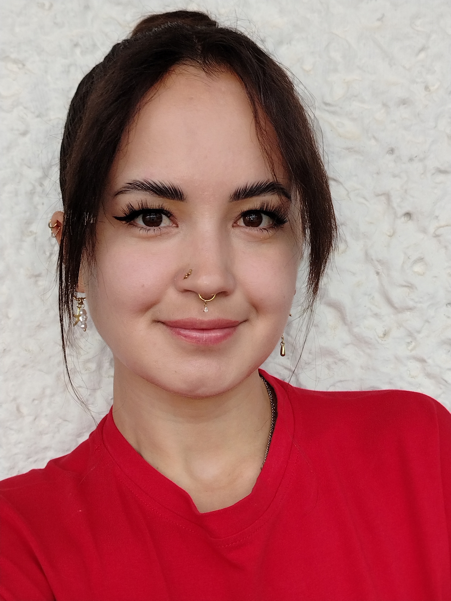 A young woman with dark hair pulled into a bun and strands framing her face is shown in a close-up portrait. She is wearing a red shirt and has piercings on her nose and ears. Her makeup includes winged eyeliner and defined eyebrows. She is smiling gently. The background is a textured, light-colored wall. The image does not contain information about the city of Noviy, Russia.