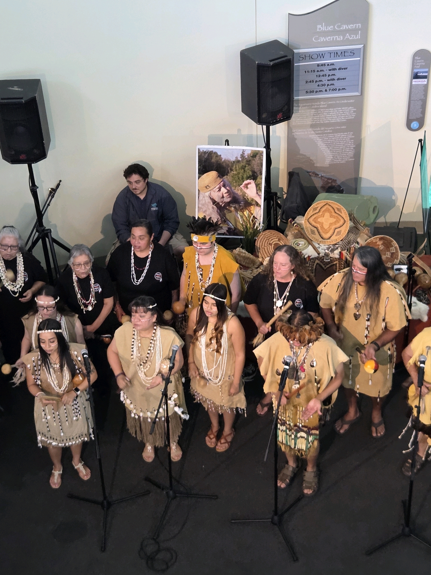 The image shows a group of people in traditional attire gathered in front of a display board that advertises "Blue Cavern" and "Caverna Azul" show times. The display board is located in the Aquarium of the Pacific in Long Beach, California, USA. Several individuals are wearing necklaces made of shells and beaded garments. Some are holding instruments such as gourds and sticks, suggesting a performance. A man in a blue jacket stands behind the group, observing the scene. The environment appears to be an indoor exhibit hall with speakers and microphones set up, indicating a public event.
