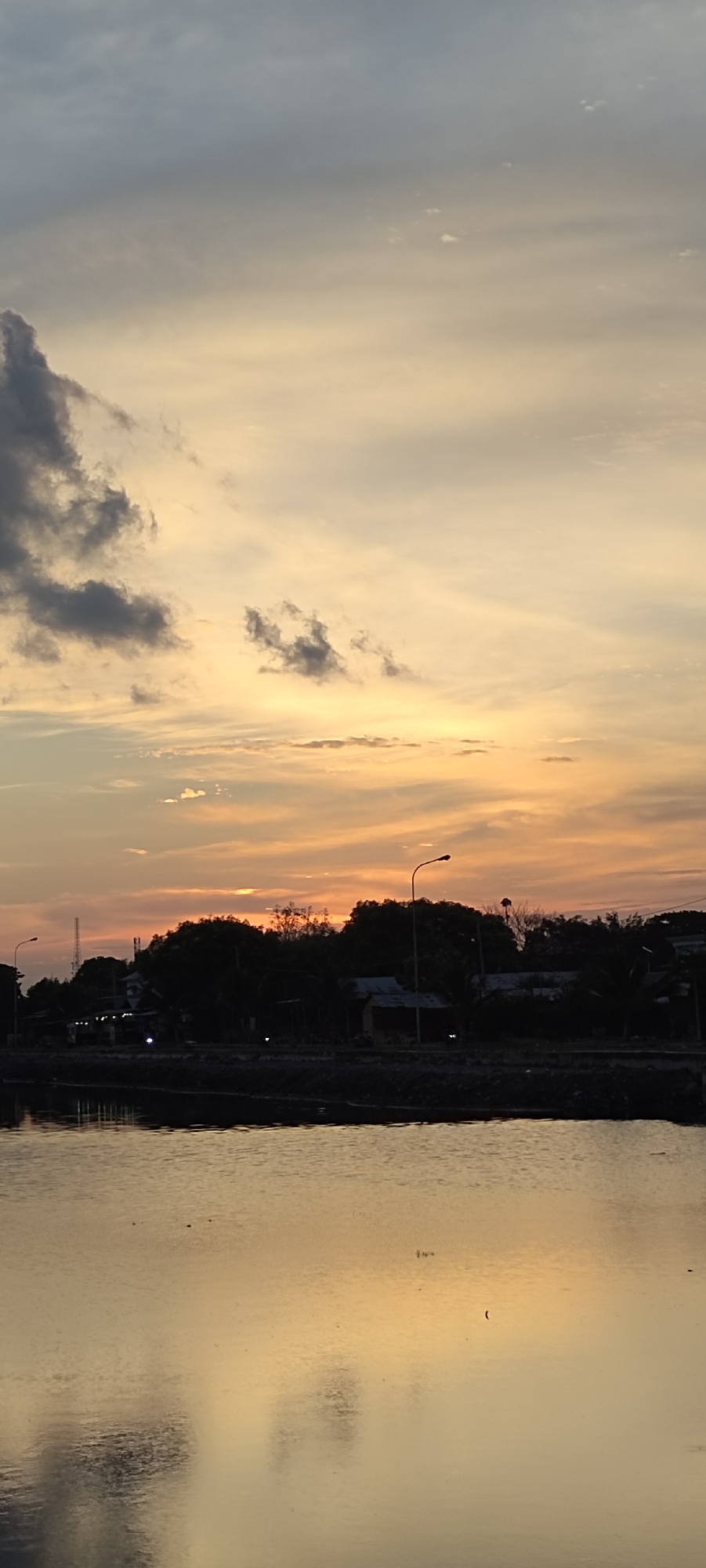 This is a vertical, full-frame shot of a serene waterfront at sunset, likely in Lhokseumawe, Indonesia. The sky is a gradient of soft oranges and yellows, with wisps of clouds tinged with grey and white. In the distance, a dark silhouette of trees and structures lines the horizon, punctuated by a streetlamp with a curved pole. The water in the foreground reflects the warm hues of the sky, creating a shimmering, textured surface. A few small, dark shapes are visible on the water, possibly birds or debris. The overall atmosphere is peaceful and tranquil, characteristic of a coastal sunset.