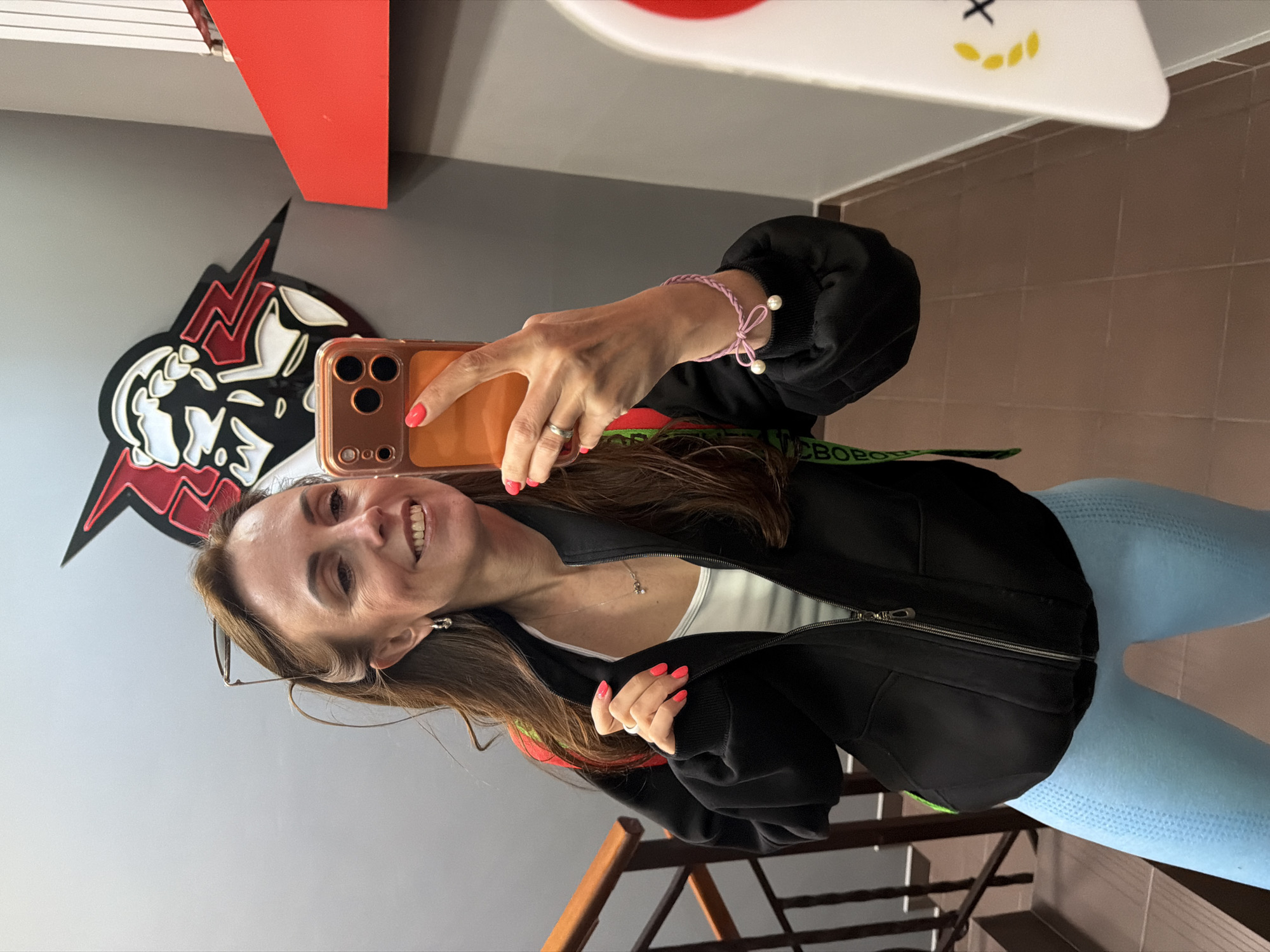 A smiling woman is captured taking a mirror selfie indoors, holding a smartphone with an orange case in her right hand. Her nails are painted a bright coral color, and she wears a silver ring on her finger, along with a pink braided bracelet adorned with two small white pearls on her right wrist. Her long, straight brown hair flows down, and eyeglasses are perched on her head. She is dressed in a black zipped jacket over a white top and light blue, textured leggings. A silver necklace with a small pendant is visible around her neck.
The background features a gray wall with a large, stylized red and white wall decor resembling the KFC Colonel Sanders logo, but with a more aggressive, lightning-bolt-like design. Above it, a red shelf or panel is visible. To the right, a section of brown tiled wall is seen, along with a wooden staircase and railing. A white surface with a yellow smiley face and a black 'X' is partially visible at the top. A green strap, possibly from a bag, crosses her body, with the partially visible Russian text "СВОБОРО" (SVOBORO) printed on it. The woman's cheerful expression suggests a pleasant mood. The indoor setting, with the distinctive logo, indicates a commercial or public space, possibly a fast-food restaurant in Birobidzhan, Russia.