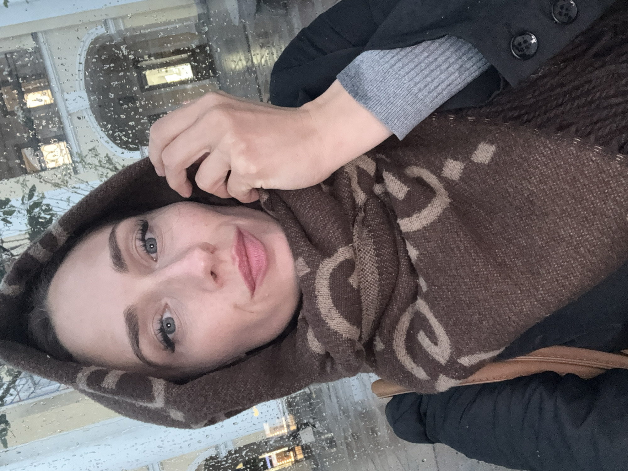 A woman with blue eyes, dark eyebrows, and red lipstick is taking a selfie from behind a window with raindrops on it. She is wearing a brown hooded scarf with a repeating tan pattern, a black jacket, and a grey sweater peeking out from underneath. Her right hand is adjusting the scarf near her face. The background shows an arched entryway to a building with lit windows. The scene is consistent with a location in Saint Petersburg, Russia, where such architecture and weather are common.