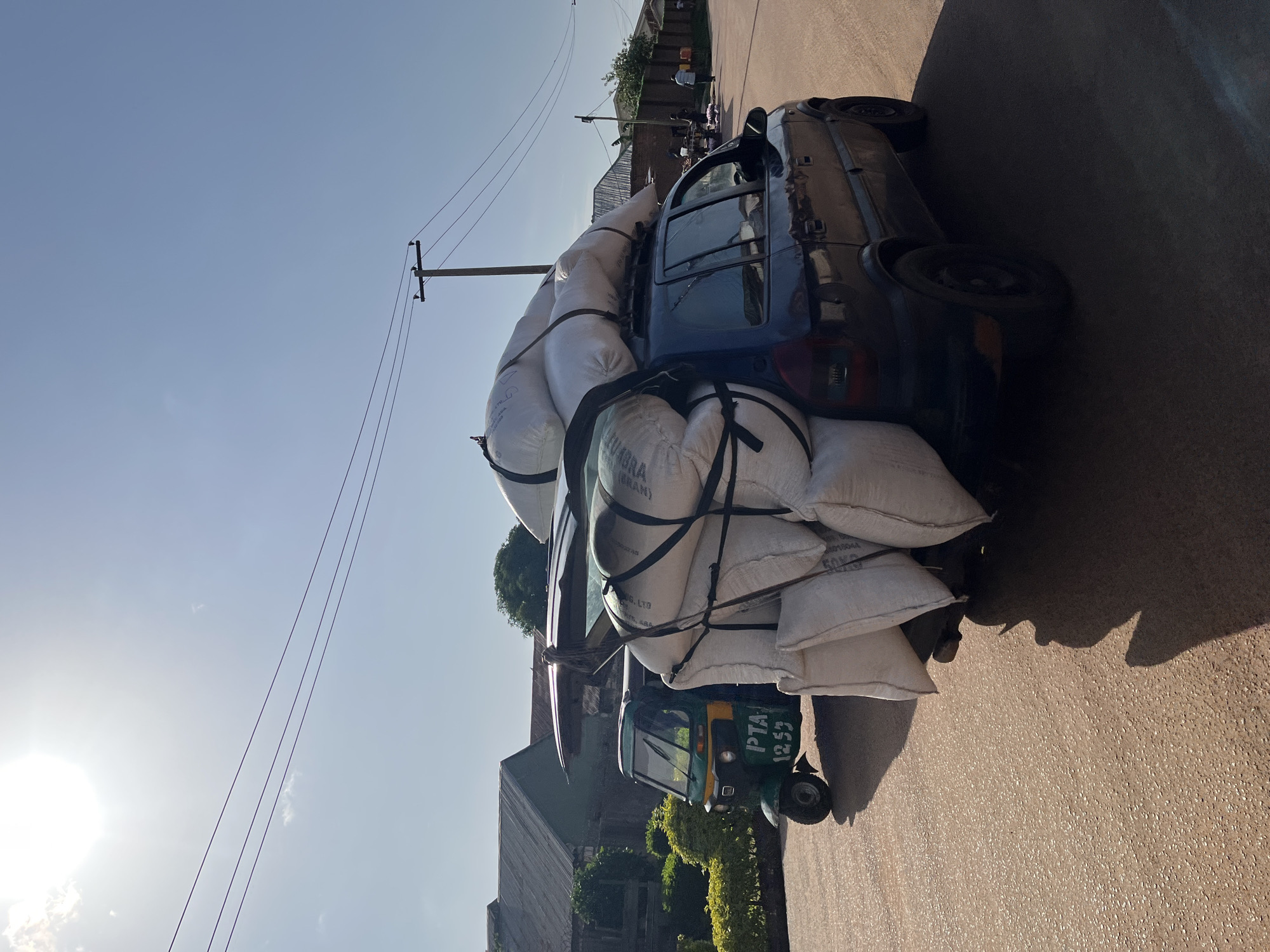 The image depicts a blue hatchback car heavily laden with large white sacks of goods, secured with black straps to the roof rack and extending over the rear of the vehicle. The car is parked on a paved road. To the left of the car is a green and yellow three-wheeled auto-rickshaw, also known as an "okada" or "keke napep," with "PTA 1253" visible on its side. In the background, there are buildings with corrugated iron roofs and some vegetation. Several utility poles and overhead wires are visible against a clear blue sky with a bright sun casting a shadow of the car and auto-rickshaw onto the road. A few people are seen in the distance, near a roadside market area. The scene appears to be in Jos, Nigeria.