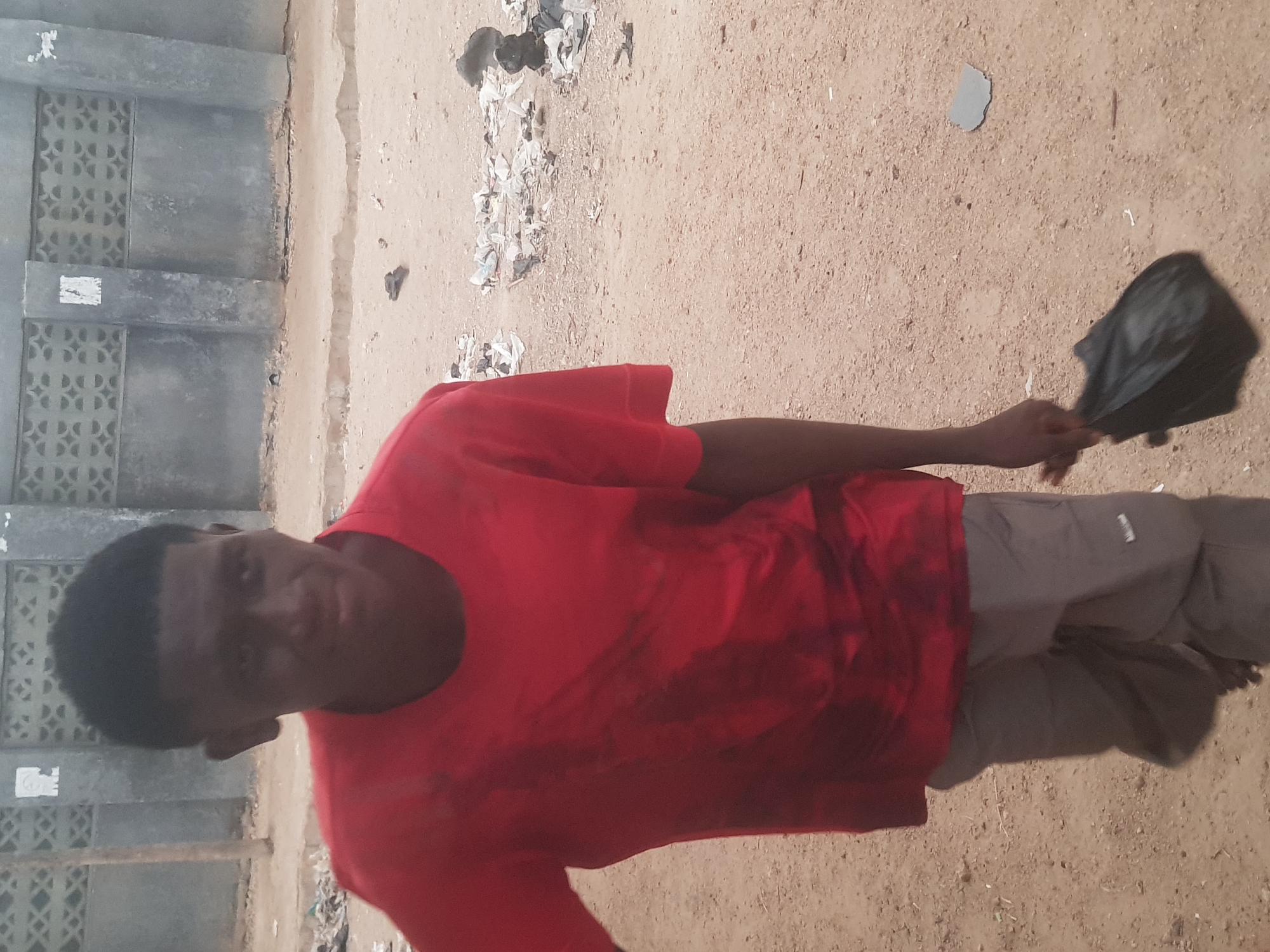 A man wearing a red shirt and gray pants stands on a sandy surface. He holds a black bag in his right hand. To the left of the man is a concrete building with decorative openings. Scattered trash is on the ground. The location is Dutsinma A, Nigeria.