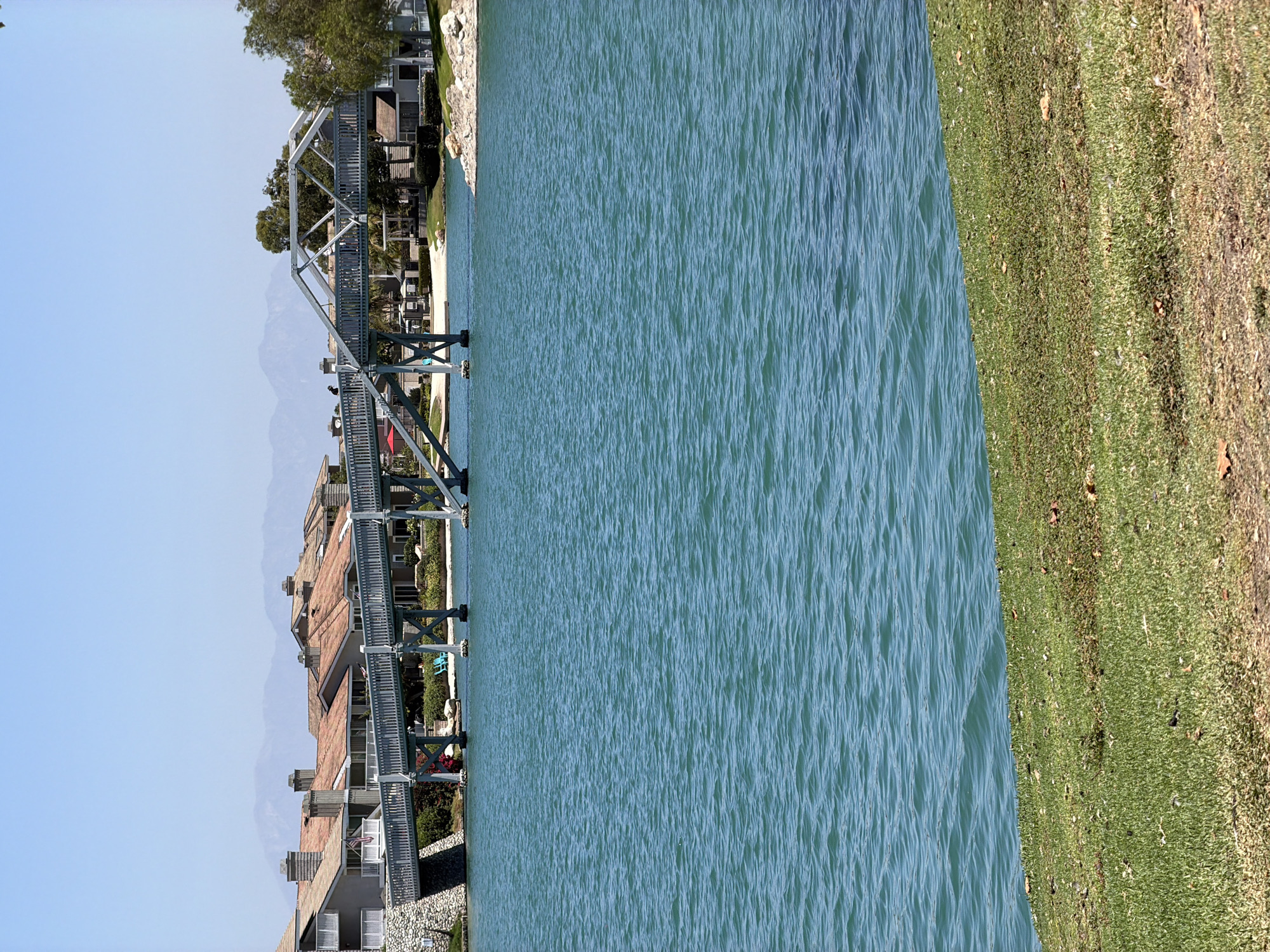 This is a vertical, medium-shot, outdoor photograph of a pedestrian bridge over a body of water, likely a lake, in Irvine, California, USA. The bridge, constructed of gray metal and wood, spans across the water with a complex, geometric design. There are a few people visible on the bridge, appearing to be walking. On the far side of the lake, behind the bridge, are residential buildings with terracotta tile roofs, indicative of a planned community. Beyond the buildings, hazy mountains are visible on the horizon under a clear blue sky. In the foreground, a grassy embankment slopes down towards the water. The water itself is a vibrant turquoise color, with small ripples on its surface, suggesting a calm day. The scene is brightly lit by sunlight.