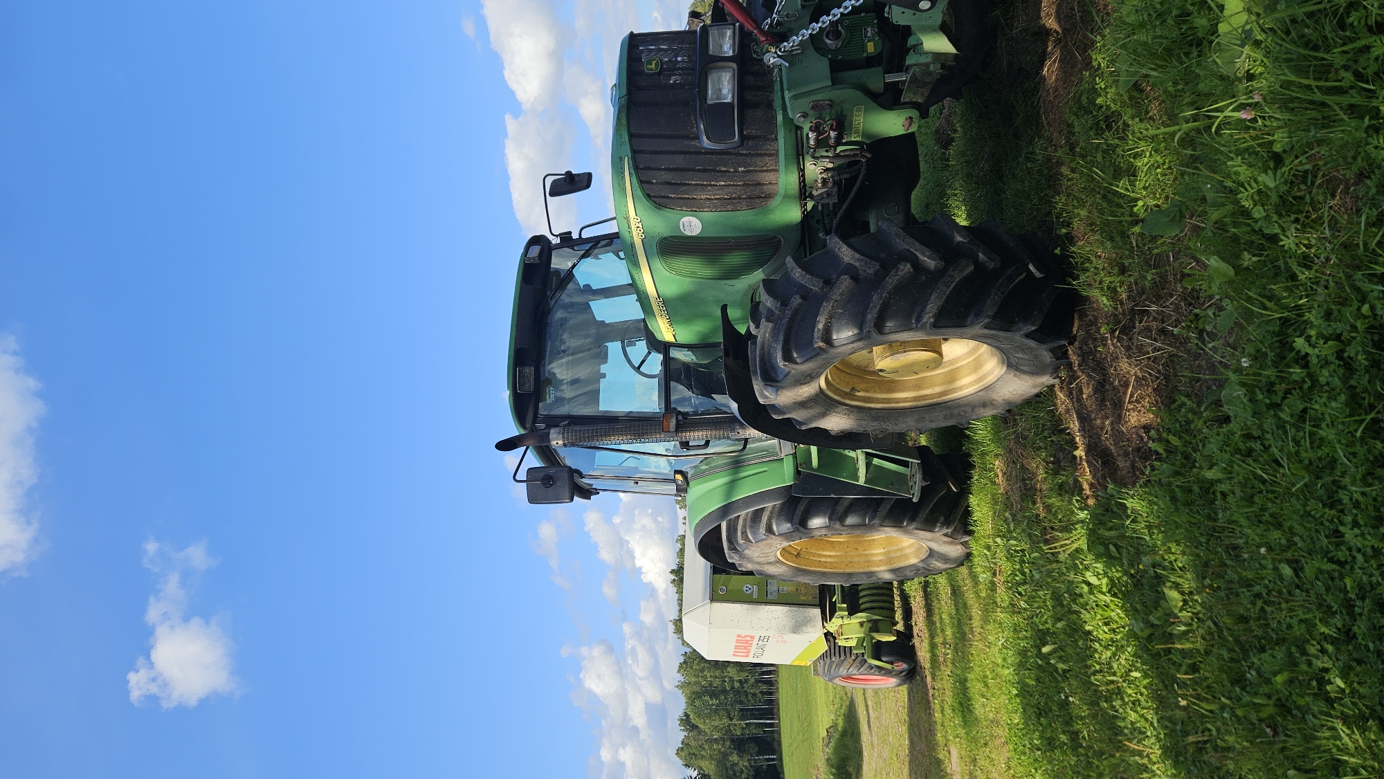 A green John Deere tractor, model 6620, is parked in a grassy field in Kapūņi, Latvia. The tractor is attached to a white Claas Rollant 255 baler. The sky is blue with scattered white clouds. Trees line the horizon in the background. The scene appears to be set in a rural agricultural area.