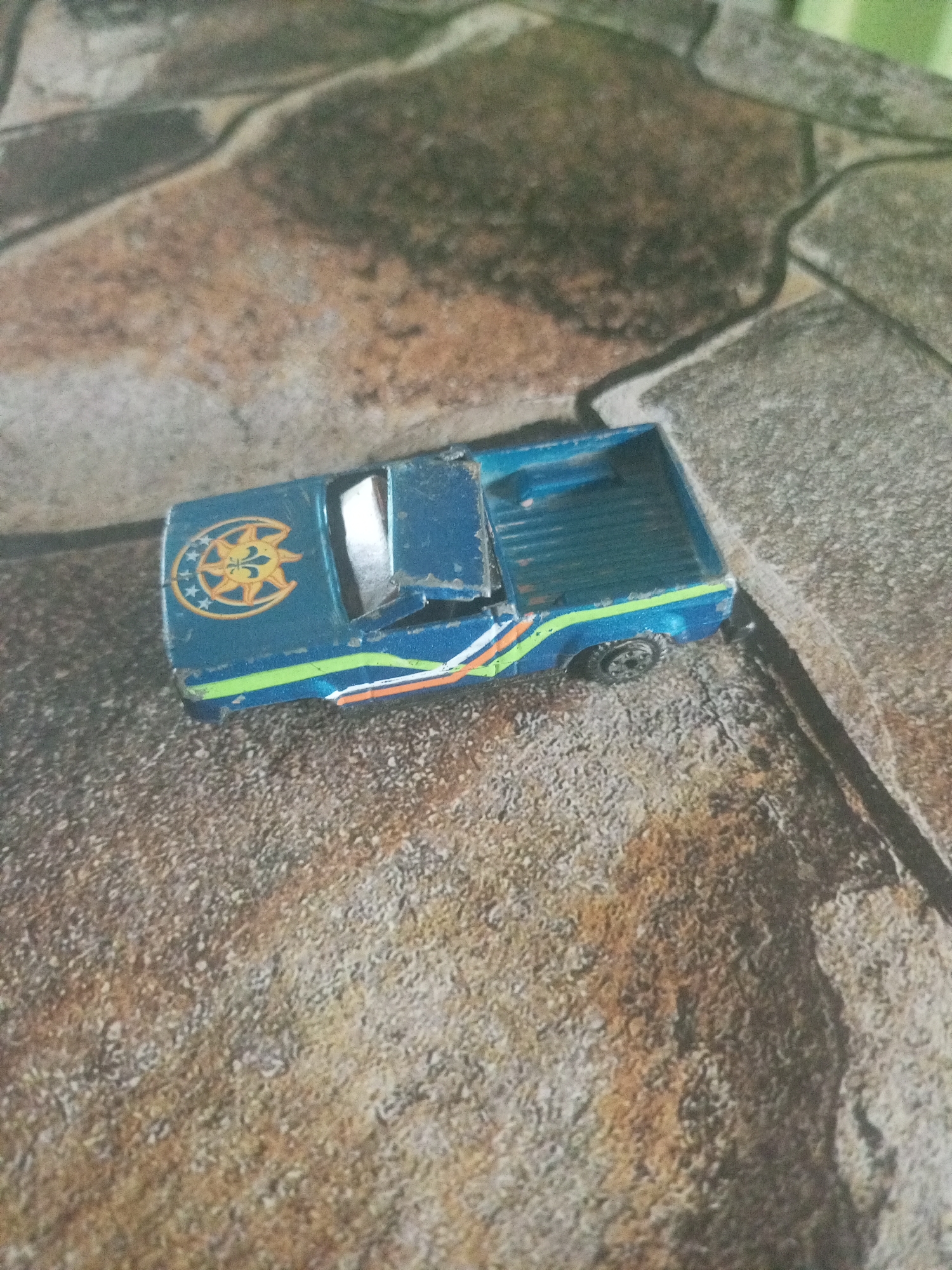 A blue, toy pickup truck rests on a surface that mimics stone tile. The truck is distressed, with scratches and a damaged windshield. It has a logo of a sun with an eye in the center, surrounded by moons and stars on the hood. Orange and green stripes adorn its side. The truck sits at an angle relative to the viewer. No people are visible. The location is not discernible as Juan Griego, Venezuela based on the image.