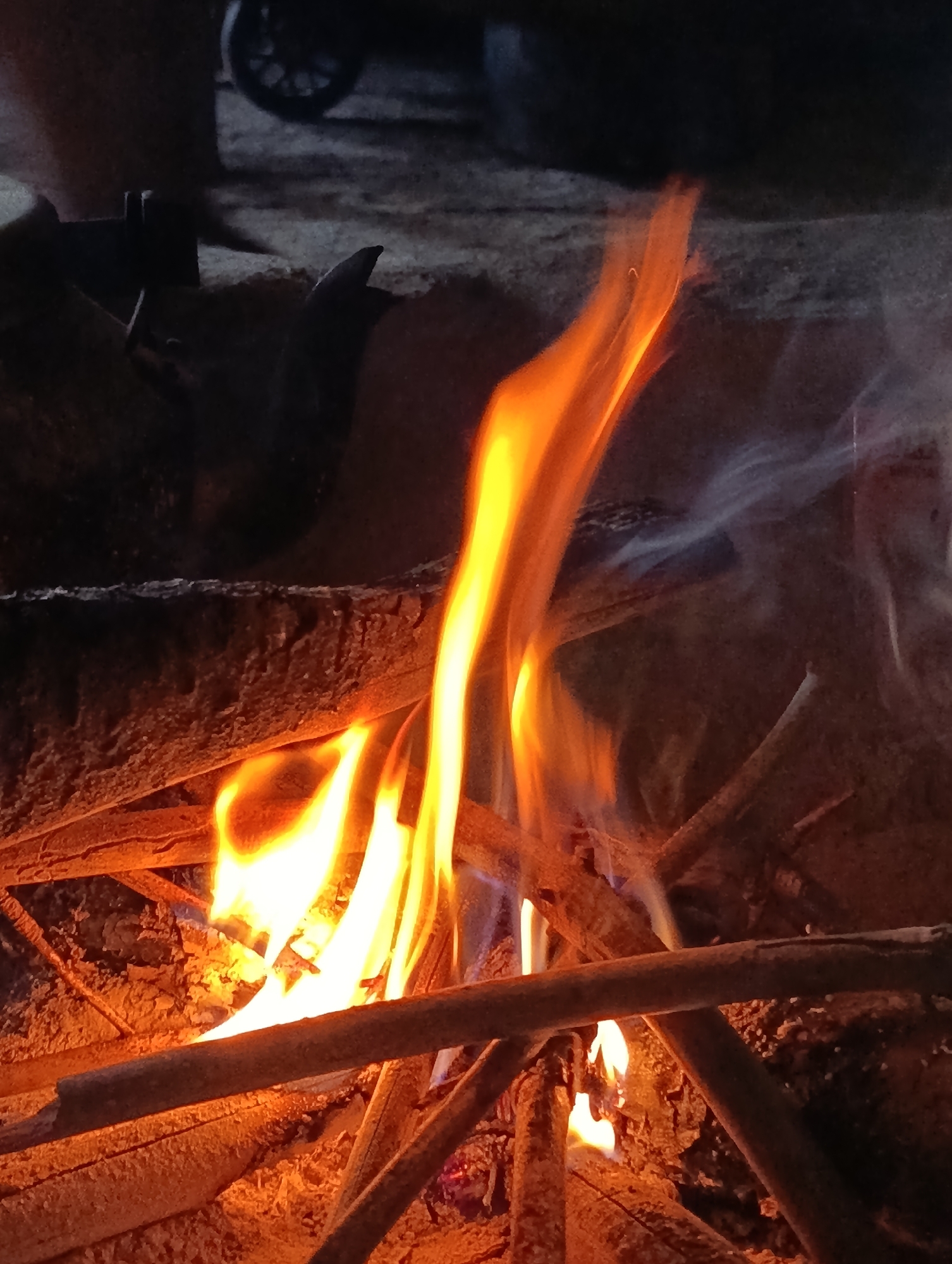 A close-up, low-angle shot captures a bonfire in Vashisht, India. The fire is composed of several branches and logs, with bright orange and yellow flames licking upwards. Wisps of white smoke rise and curl from the fire, blending with the dark, out-of-focus background. In the upper left quadrant, a dark, blurred object with a circular element, possibly a wheel, is visible. The ground beneath the fire appears to be composed of dirt and small stones. The overall scene is dark and dimly lit, emphasizing the glow of the flames.