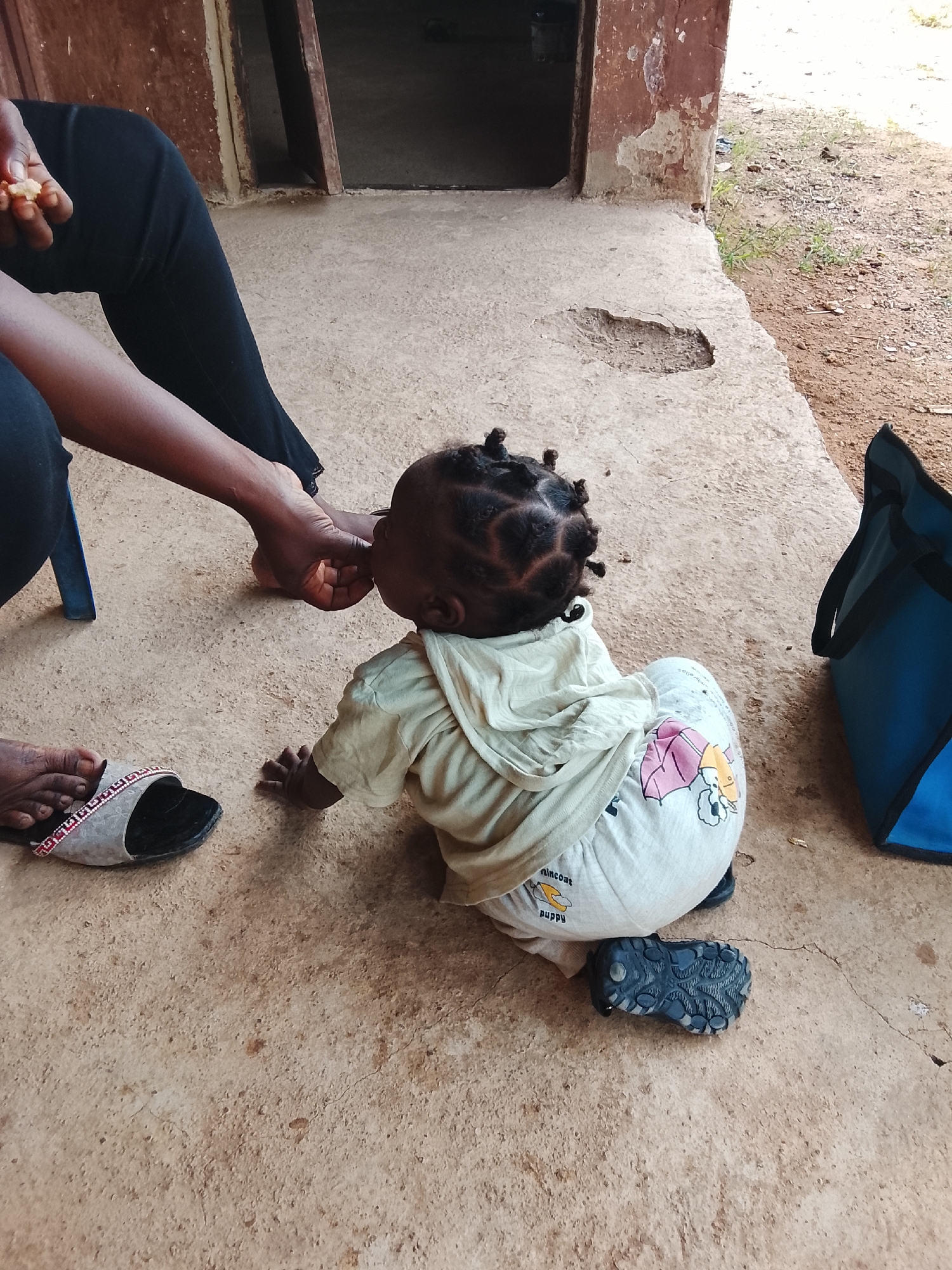 A medium shot captures a baby with black braided hair sitting on a beige, cracked concrete surface. The baby is wearing a beige hooded shirt and patterned white pants, with a dark blue shoe partially visible beneath them. An adult hand offers the baby food, while another adult, wearing black pants, sits nearby with a sandal-clad foot visible. A blue tote bag rests on the ground next to the baby. The scene is located outside a building with a doorway visible in the background. The location is Akwanga, Nigeria.