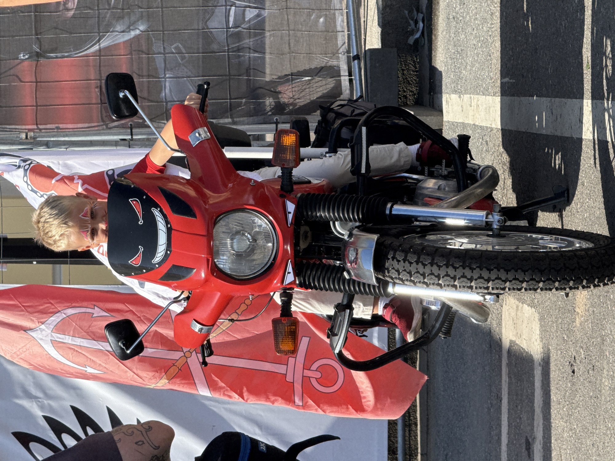 A young boy with face paint is sitting on a red motorcycle with a black windshield that has cartoonish eyes and a grin. He is wearing a white shirt and light-colored pants with red sneakers. The motorcycle has a black crash bar, chrome accents, and exposed engine components. Behind the motorcycle are large red and white flags with nautical symbols. The scene appears to be outdoors, possibly at an event, with a concrete barrier and fencing in the background. The ground is paved asphalt with white lines. The boy is holding the handlebars and looking towards the camera. The overall lighting suggests it is daytime. The location is not identifiable within the city of Saint Petersburg, Russia.