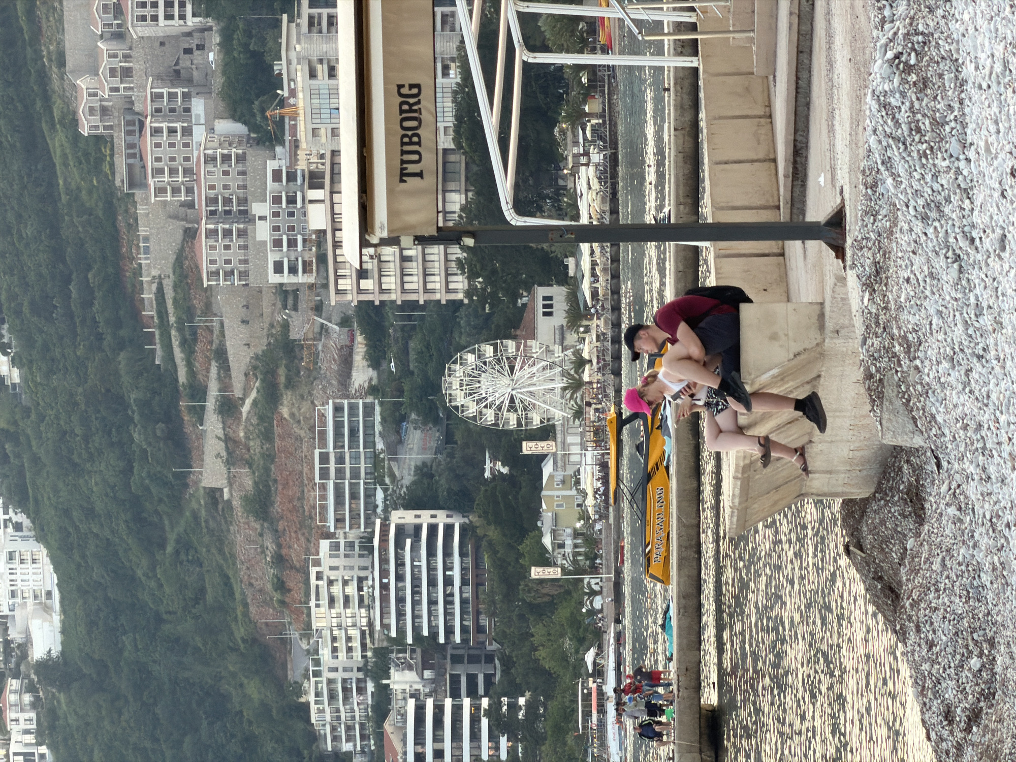 Here's a description of the image:
The image shows a view in Budva, Montenegro, likely taken from a higher vantage point overlooking the town and the Adriatic Sea.
In the foreground, there is a section of a concrete structure, possibly a pier or wall, with a beach nearby covered in pebbles. A couple is present; a man leans down towards a woman wearing a pink hat. There is a yellow "Parasailing" stand next to them. Above them is a "TUBORG" sign.
In the background, there's a Ferris wheel and various modern buildings interspersed with greenery along the hillside. The sea is visible with sunlight reflecting on the water's surface. A small crowd of people is also noticeable on the pier near the water.
