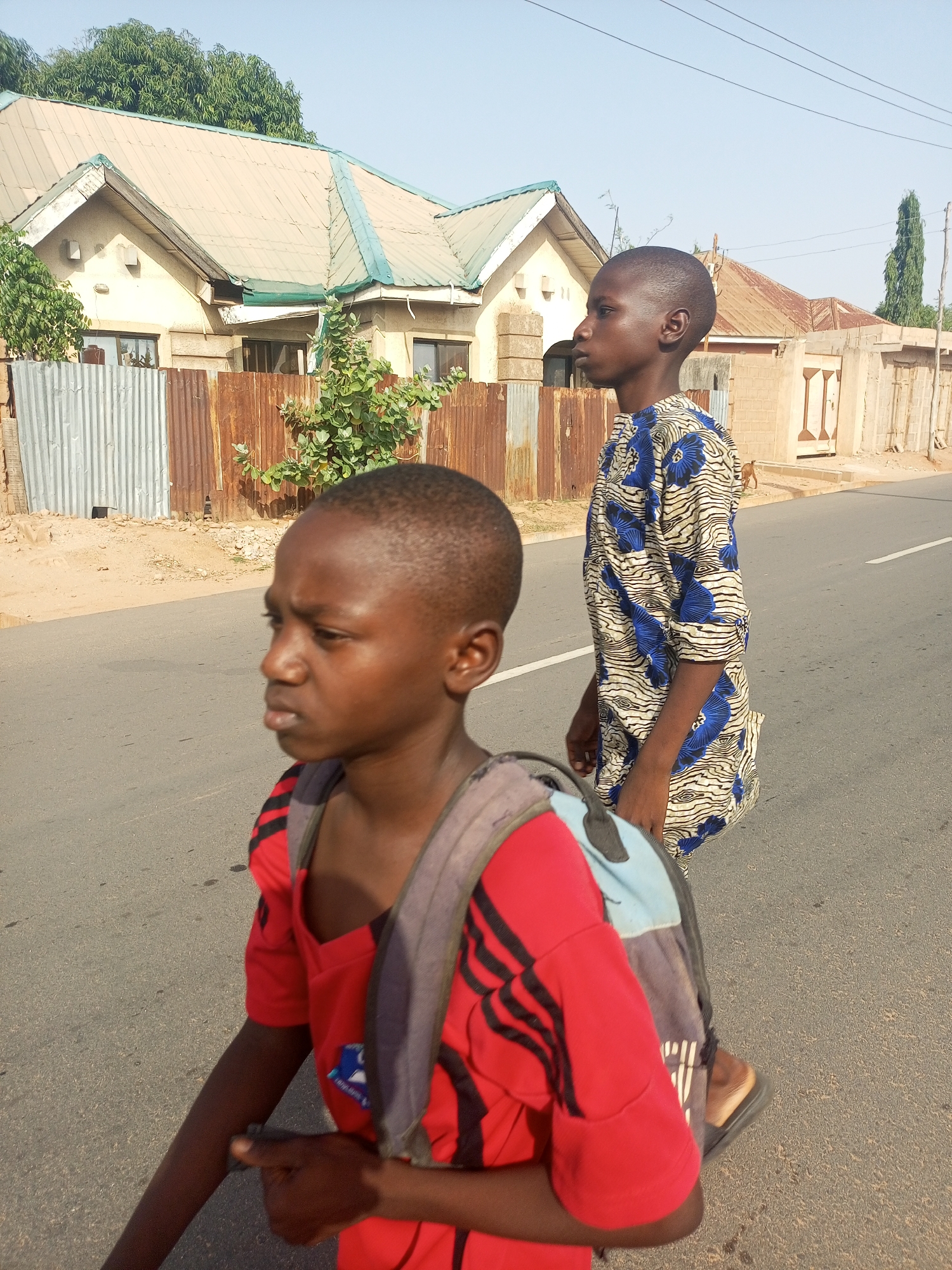 Two boys walk along a paved street. The boy in the foreground wears a red shirt with black stripes and a blue backpack. The boy behind him wears a blue and white patterned shirt. They are walking on a road in Tempure, Nigeria. Behind them, there are houses with corrugated metal fences. The houses have light-colored roofs and walls. A few trees are visible in the background. The sky is light blue.