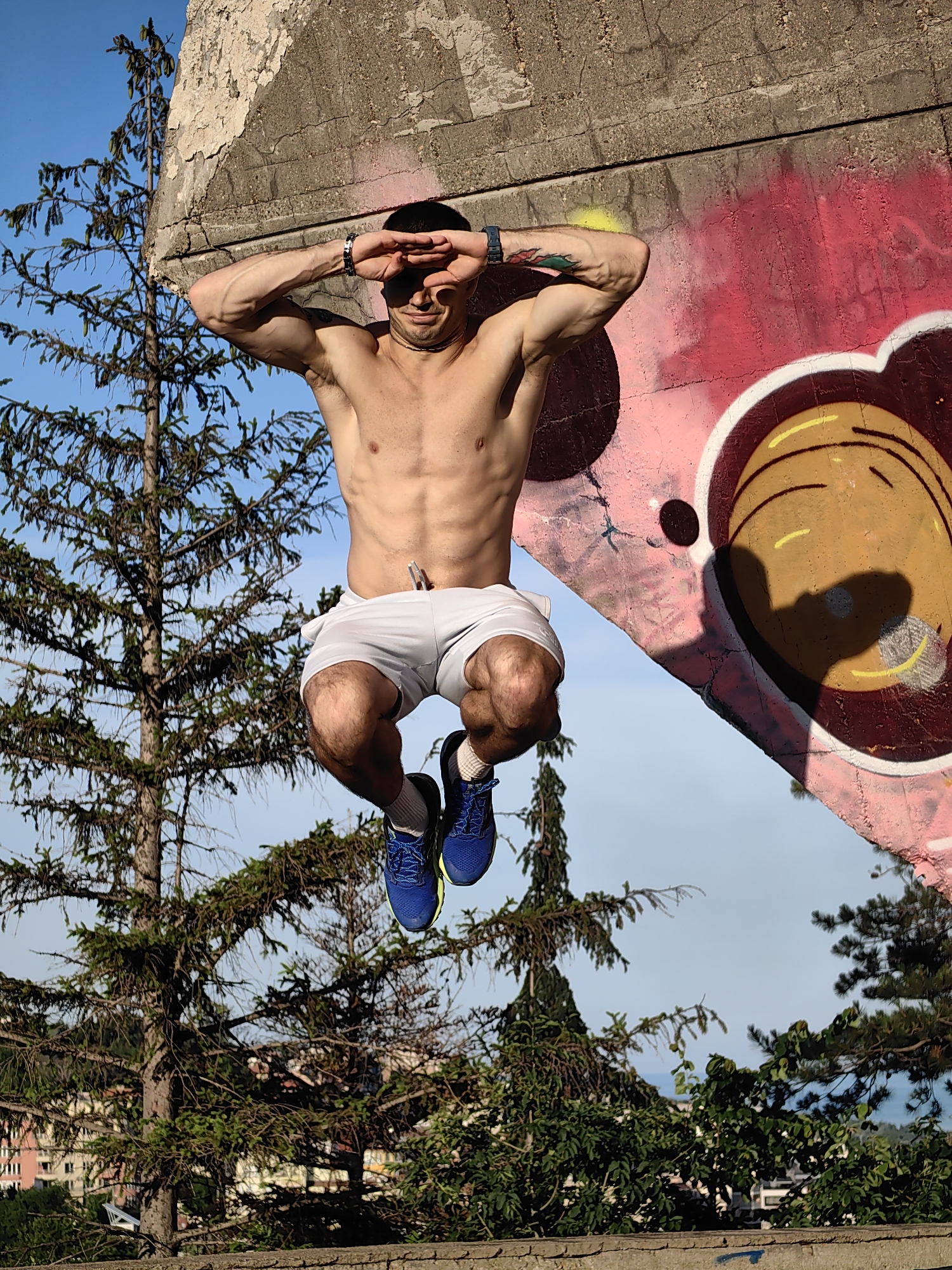 A shirtless, muscular man in gray athletic shorts and blue running shoes is captured mid-air in a jump, his arms raised and hands shielding his eyes. He is positioned in front of a concrete structure adorned with vibrant graffiti, including a large pink design with circular elements and a shadow of a face. The background reveals a clear blue sky, pine trees, and a distant view of buildings and greenery, suggesting an outdoor urban setting. This image was taken in Varna, Bulgaria, specifically near the Varna Observatory.