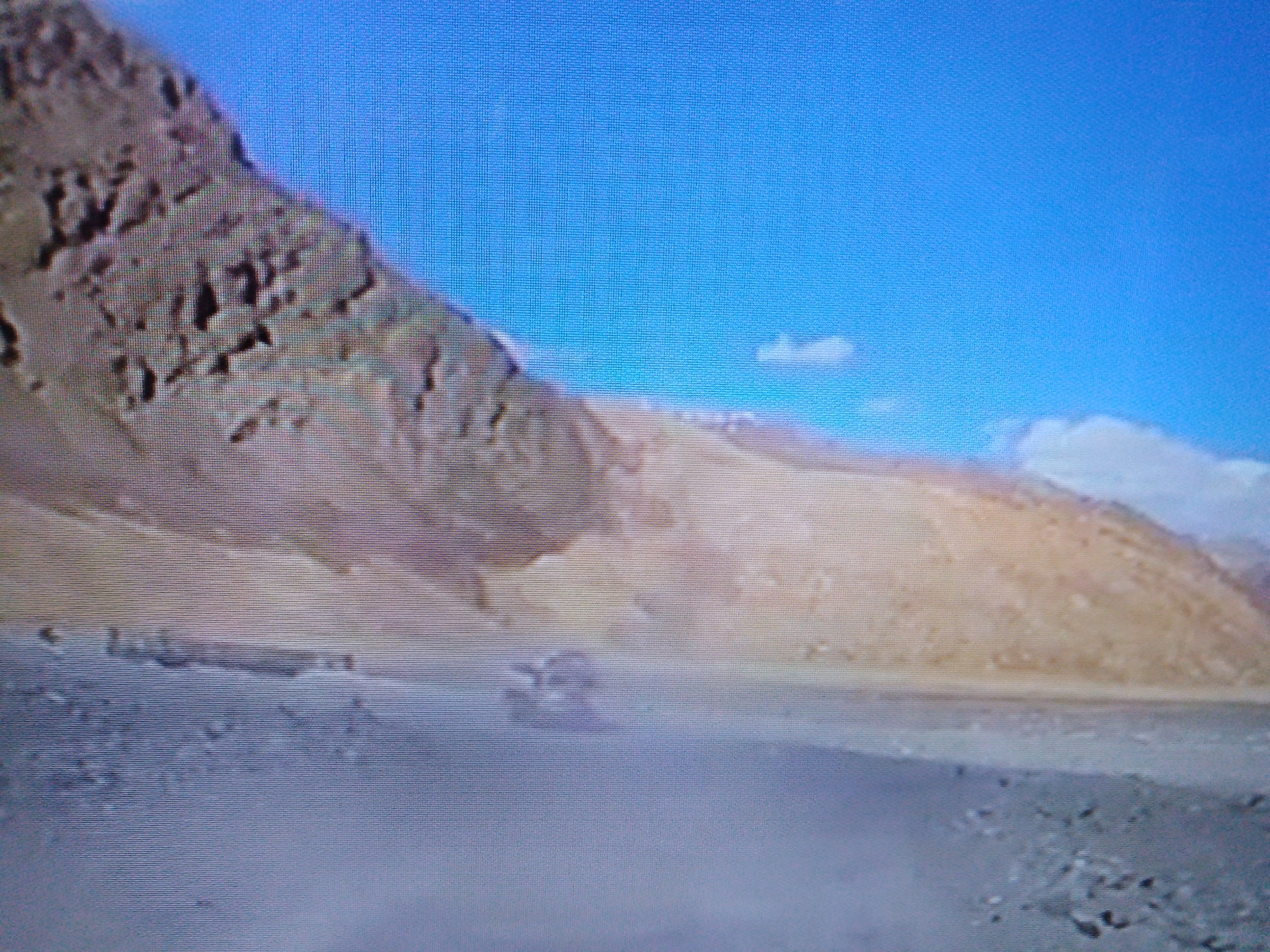 The image shows a desert landscape with a vehicle driving in the distance, kicking up dust. The terrain is arid and mountainous, with a clear blue sky above. The vehicle appears to be a rugged, off-road type, possibly an SUV or truck, and it is moving at a speed that is causing a noticeable dust cloud to trail behind it. The overall scene suggests a remote and possibly challenging environment. There is no indication that this image is related to the city of Dushanbe, Tajikistan, as there are no identifying landmarks or features present.