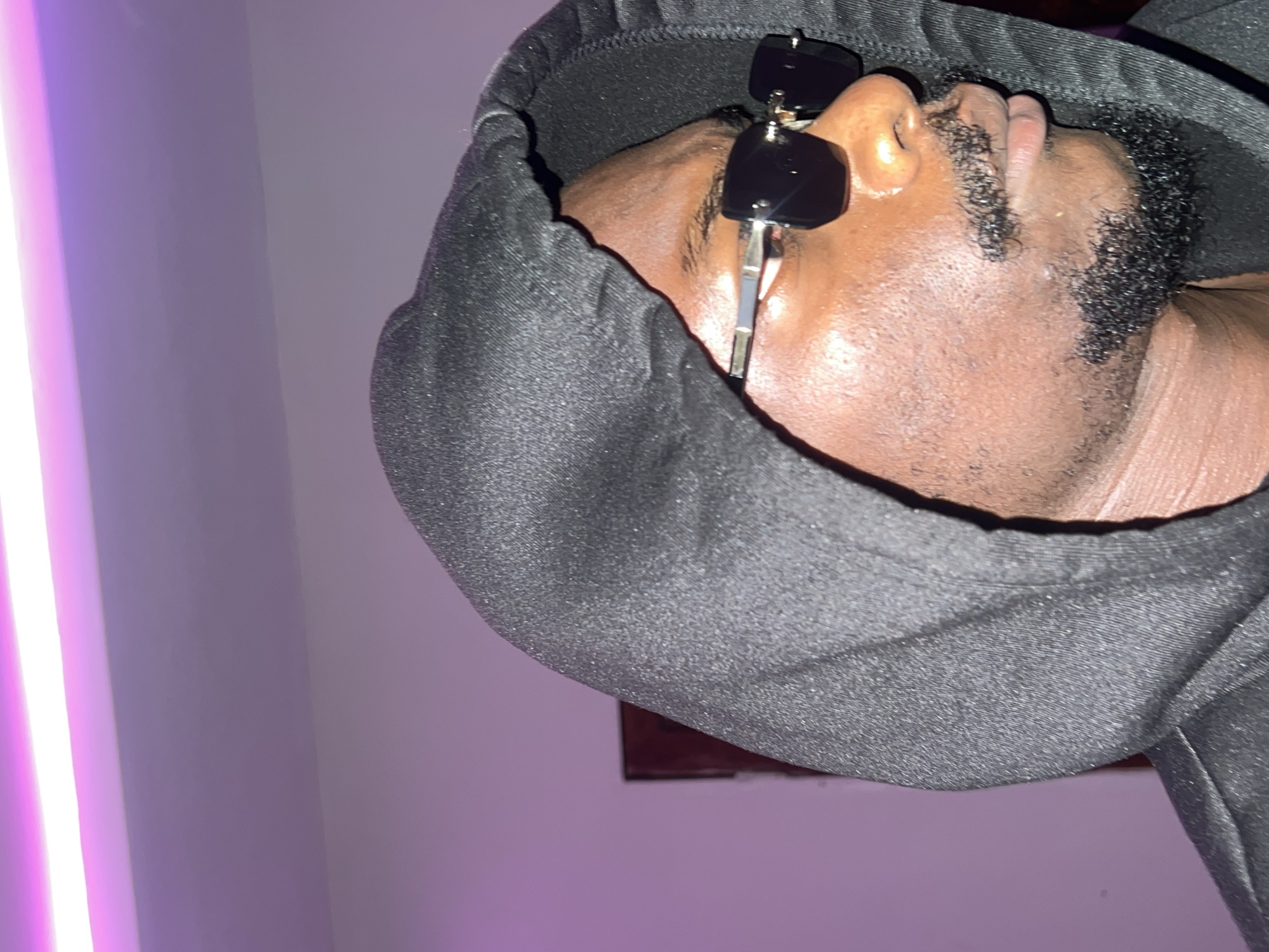 A person wearing a black hoodie and sunglasses is shown from a close-up, tilted angle. The subject appears to be a black man with a short, trimmed beard. The background includes a wall illuminated by a vertical light source, possibly a neon tube, casting a pinkish-purple hue. In the lower part, a shadow suggests the presence of a framed object. Specific details to confirm the location in Lagos, Nigeria, are not visible in the image.