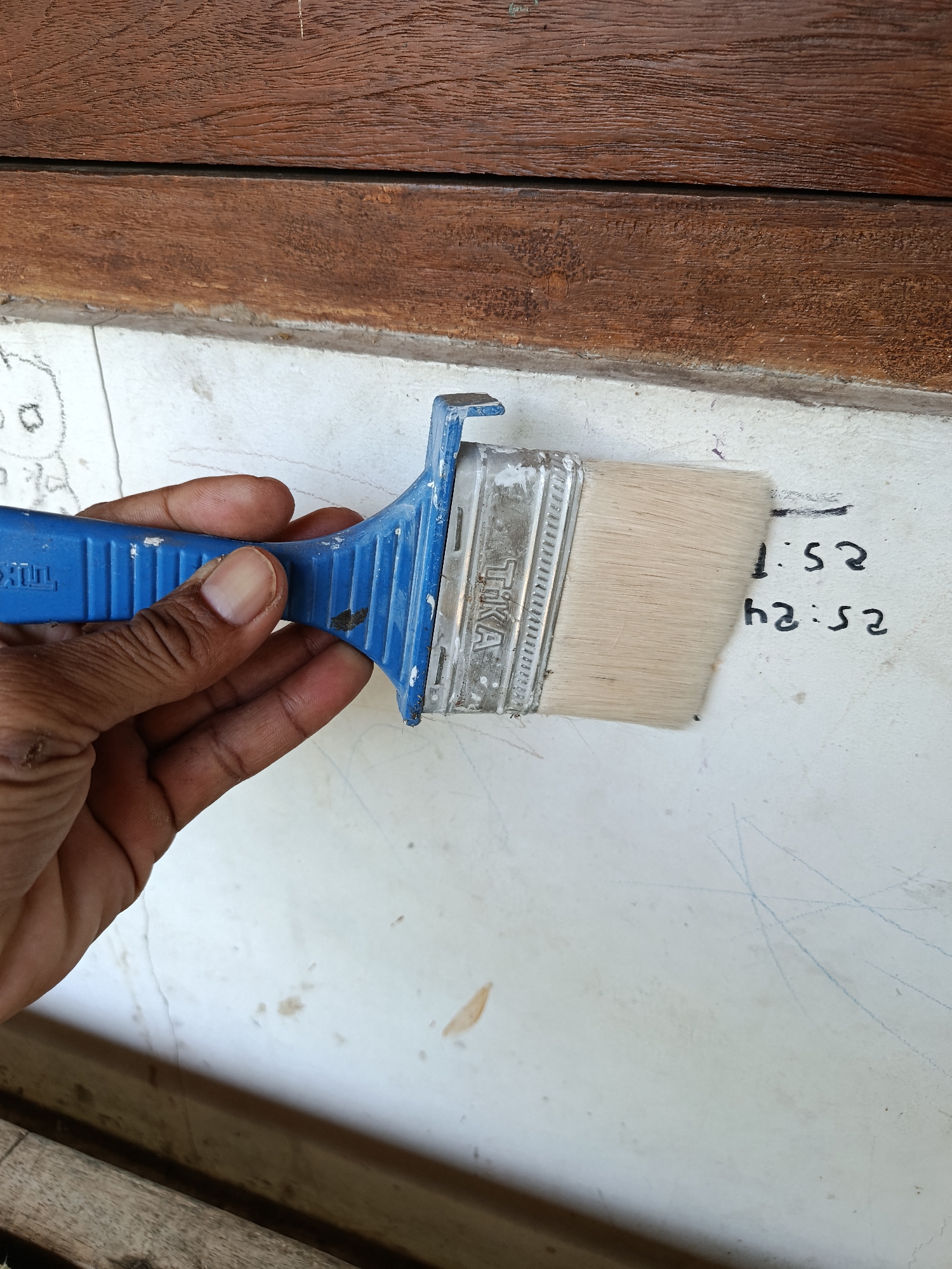 A hand holds a blue-handled paintbrush with a metal ferrule and light-colored bristles, positioned against a white wall with faint blue scribbles and some brown smudges. The wall is bordered by two horizontal wooden planks, the upper one appearing darker and more weathered than the lower one. The scene is likely located outdoors, given the natural lighting and the rustic textures of the wood and wall. The presence of the paintbrush suggests an activity related to painting or preparation for painting. There are no visible indications of the specific location within Kemiri, Indonesia.