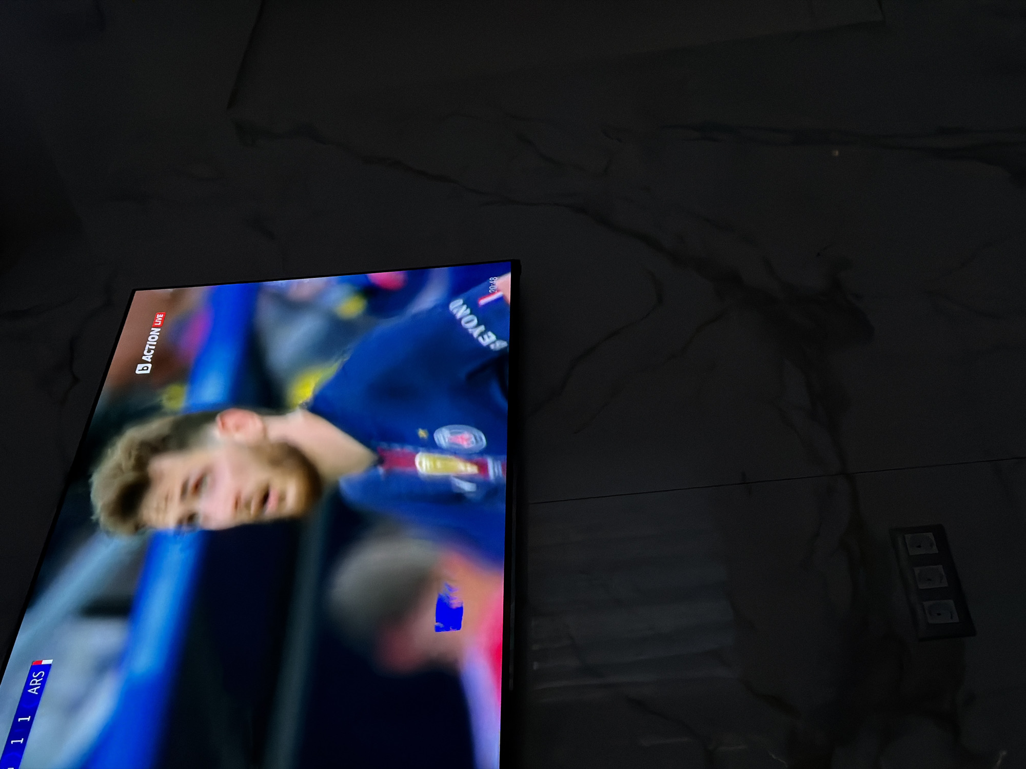 A television screen displays a soccer game between ARS and another team. The score is 1-1.
The television is positioned on a marble or tile wall.
The location, Asenovgrad, Bulgaria, is not indicated in the image.