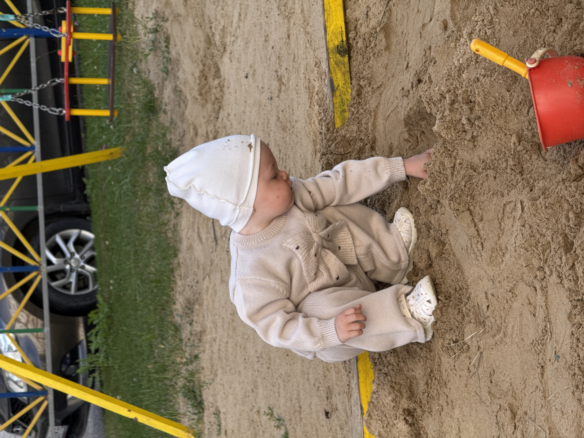 A young child, wearing a beige knitted jumpsuit and a white hat, is playing in a sandbox. The child is kneeling and digging in the sand with their hands. A red plastic bucket and a yellow plastic shovel are visible in the foreground. In the background, a swing set and a car are partially visible. The sandbox appears to be located in an outdoor play area, possibly a park or a backyard, in the city of Vladimir, Russia. The lighting suggests it is daytime.