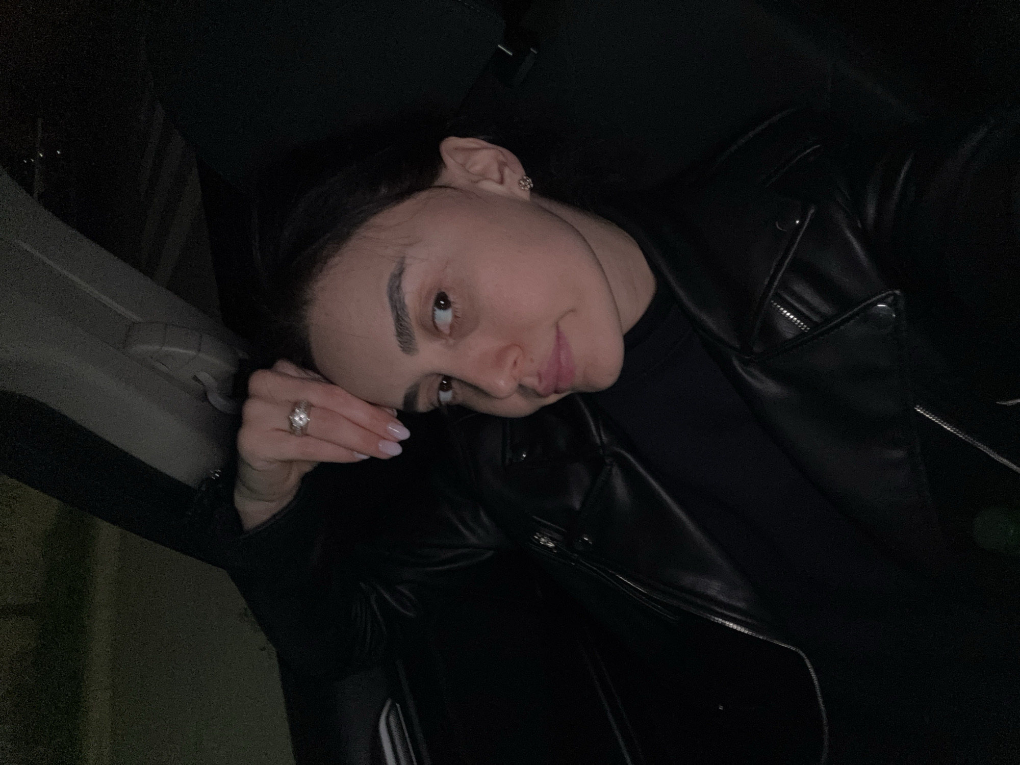 A young woman is lying in a car wearing a black leather jacket and a dark top. She is resting her head on her hand, on which she has a large ring. She is looking towards the camera. The background is mostly dark and indistinct, though a car door frame is visible. The setting appears to be at night, but the exact location within Tbilisi, Georgia cannot be determined from the information available in this image.