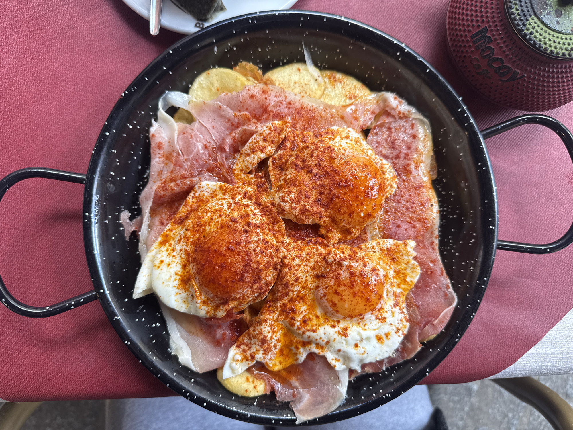 A top-down, close-up shot shows a black paella pan with fried eggs, ham, and sliced potatoes, liberally sprinkled with paprika, resting on a table with a red and white striped tablecloth. A small white plate with utensils is partially visible in the upper left, and a red container with the word "Mocay" on its side is in the upper right. The dish appears to be a serving of "huevos rotos," a typical Spanish dish, likely photographed in Madrid, Spain, given the food's popularity in the area. The focus is solely on the dish and the immediate surrounding table setting, with no people visible in the frame.