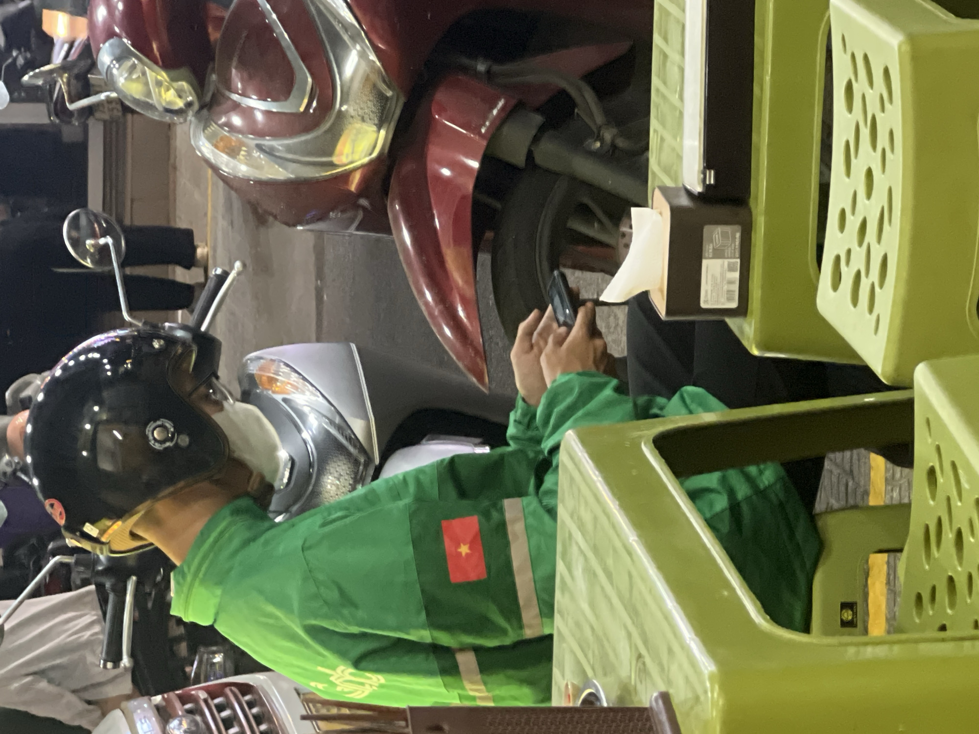 In Đà Nẵng, Vietnam, an individual wearing a green jacket with the Vietnamese flag patch and a motorcycle helmet uses a smartphone near a motorcycle, adjacent to green plastic crates and a tissue dispenser.