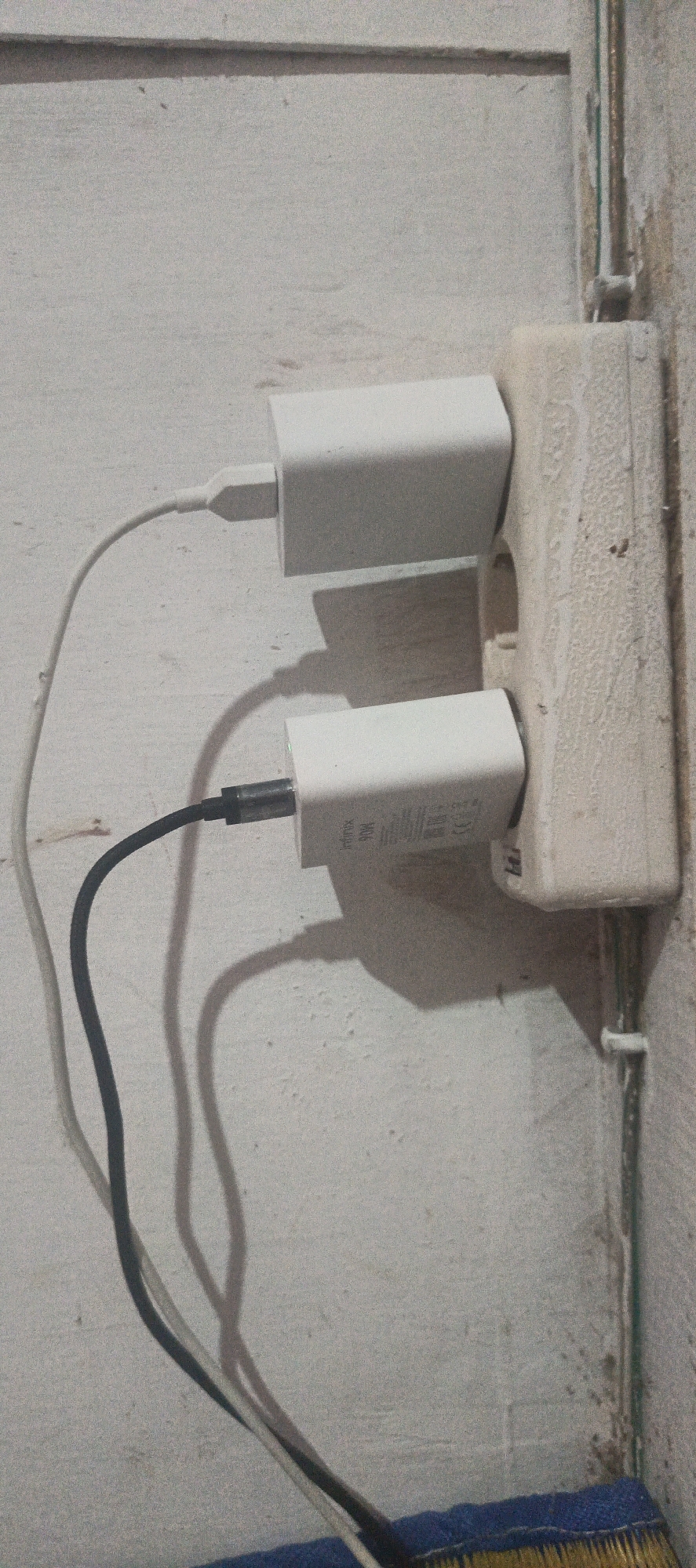 In Cinta Dame, Indonesia, two white power adapters are plugged into a dual electrical outlet mounted on a textured, off-white wall. The top adapter is connected to a white charging cable, while the bottom adapter is connected to a black charging cable. The cables are visible extending downwards and to the left. The electrical outlet appears aged and has some discoloration. A portion of a broom with yellow bristles and a blue fabric cover is visible at the bottom of the image, suggesting the location is likely a room or utility space. The lighting creates shadows cast by the adapters and cables on the wall.