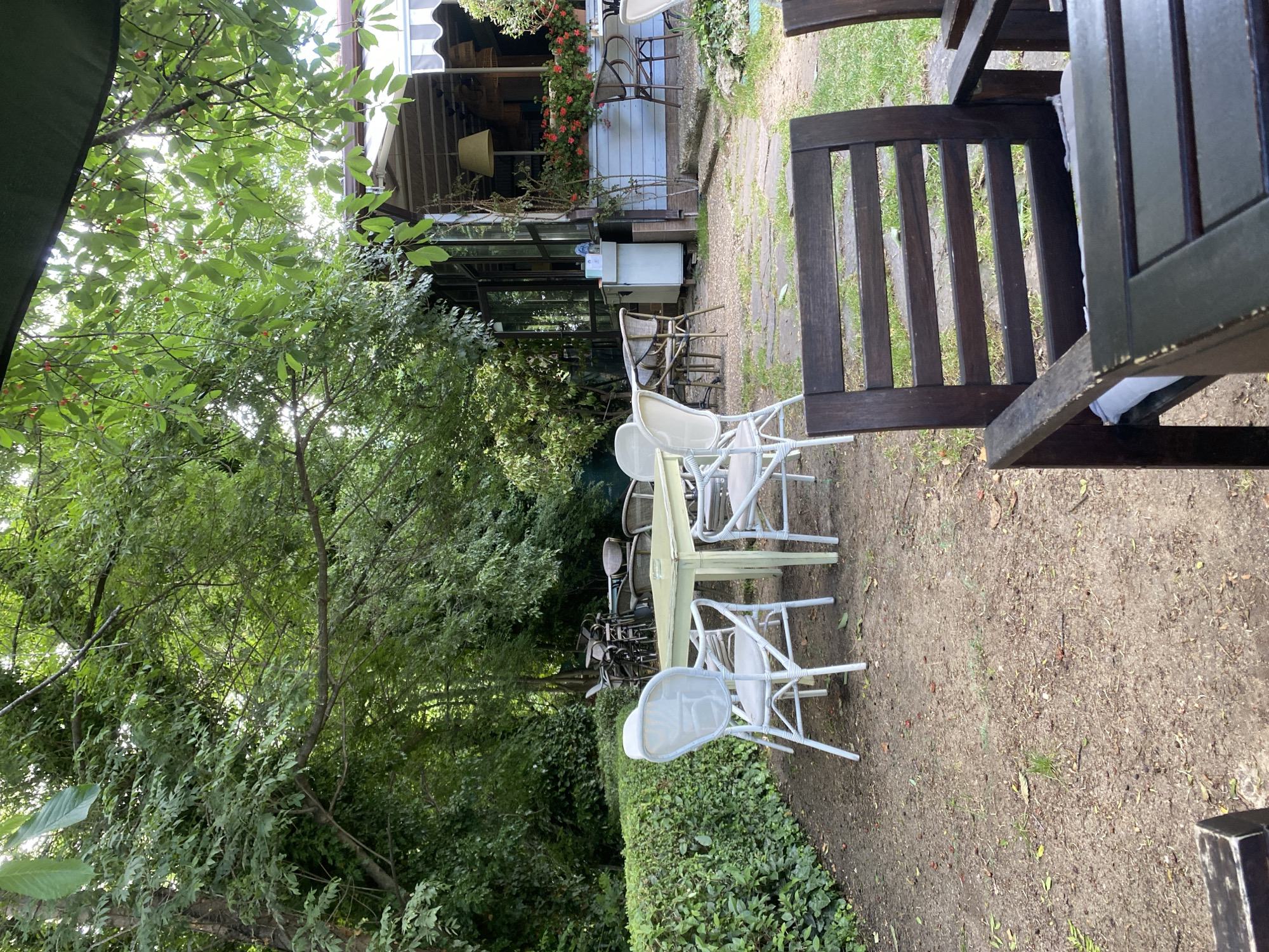 This image depicts an outdoor seating area at a cafe or restaurant, likely located in Varna, Bulgaria. The scene is set amidst lush greenery, with trees and hedges dominating the background and framing the seating arrangements. Several tables and chairs are visible, some made of dark wood and others of white metal. One table is a light green color and appears to be made of metal as well. The seating area suggests a relaxed, outdoor dining experience. The lighting suggests it is daytime, possibly late morning or afternoon, with sunlight filtering through the leaves. There are no people visible in the frame, and no specific event or activity is taking place; the scene appears to be set up for customers. The overall impression is one of a tranquil, natural setting designed for outdoor leisure.