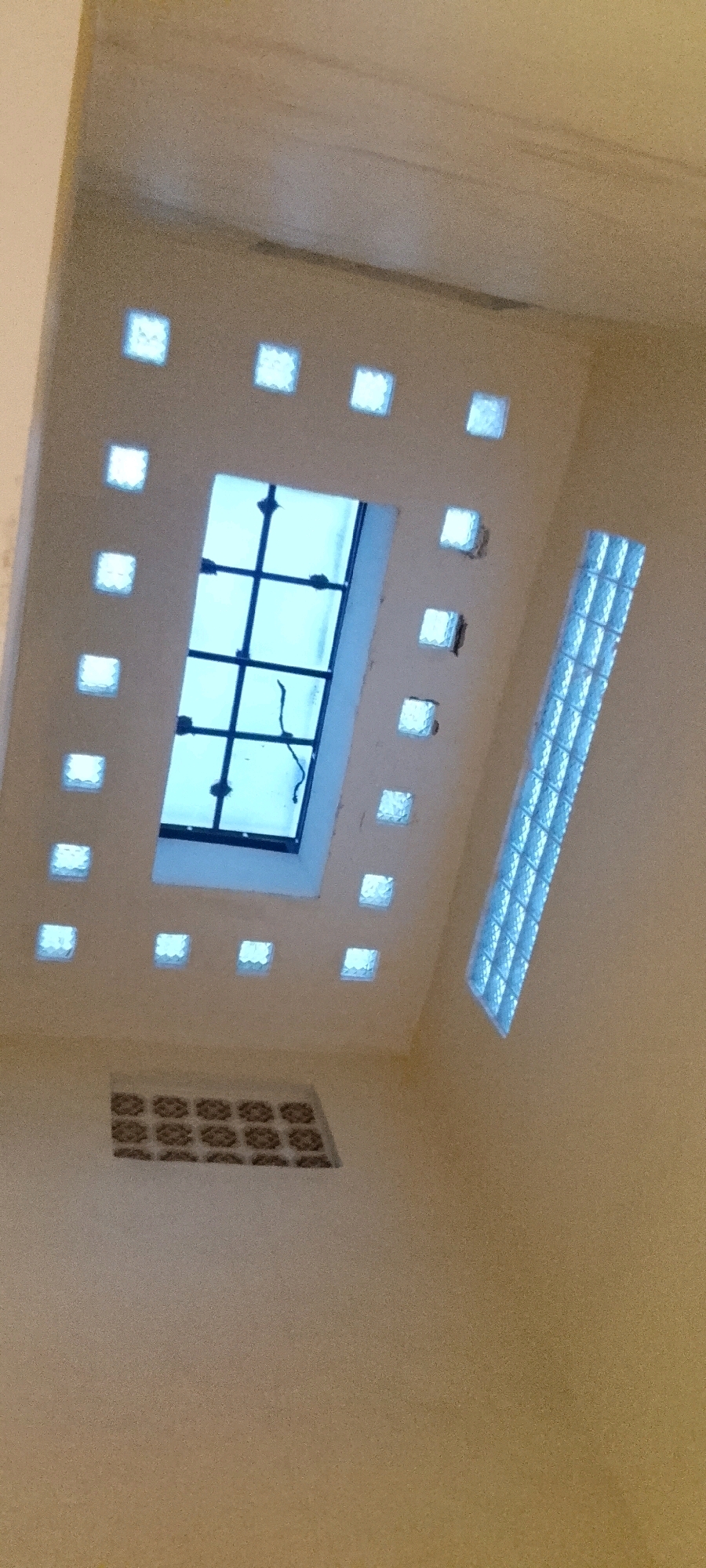 This is a high-angle interior shot of a building's ceiling in the City of Medan, Indonesia. The ceiling is predominantly a neutral, light brown color. Dominating the upper center of the image is a rectangular skylight with a dark grid pattern, letting in bright blue light. Surrounding this skylight are numerous small, square glass blocks, each emitting a soft, white light, creating a decorative border. To the right of the skylight, a vertical panel of translucent, blue glass blocks is affixed, also emitting light. In the lower portion of the image, near the center, a smaller, rectangular vent with a patterned grille is visible. The overall impression is one of artificial and natural light sources illuminating an architectural interior. There are no people or discernible actions taking place.
