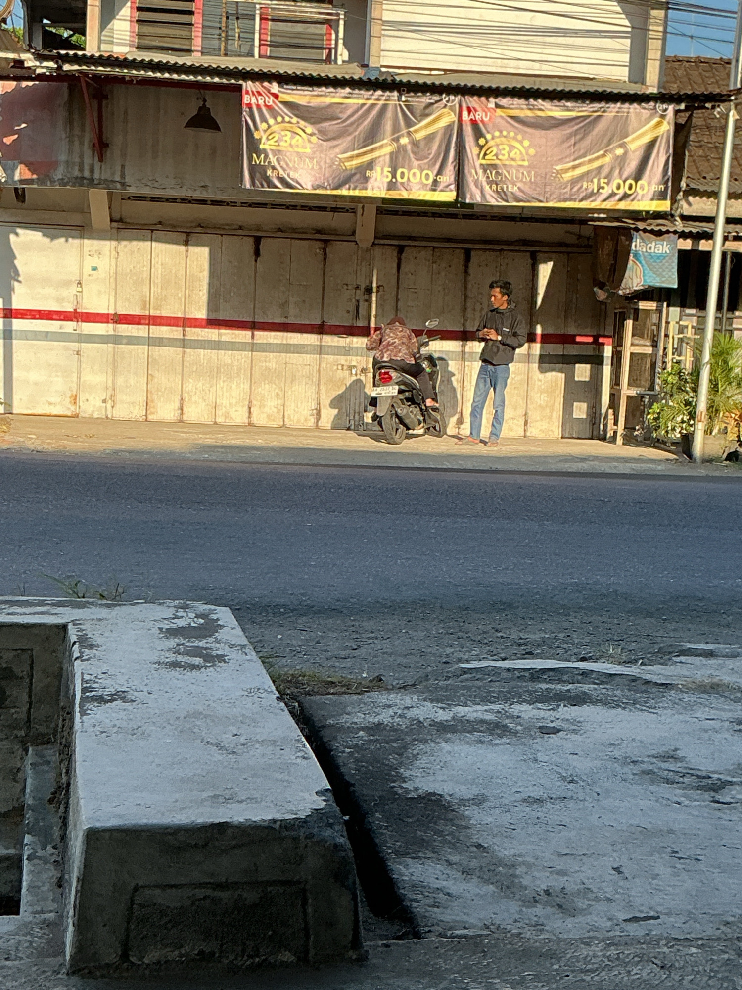 In the city of Kebonrejo, Indonesia, a street scene unfolds with two individuals and a motorcycle parked on a sidewalk in front of a building with closed metal shutters. The building features a corrugated metal roof and a large banner advertising "MAGNUM KRETEK" for RP 15,000. The banner has a gold design with imagery of cigars. A man in a black jacket and blue jeans stands near the motorcycle, appearing to be engaged in a conversation or waiting. A second person, partially obscured by the motorcycle, is seated on it, wearing a patterned shirt. The street in front is paved, and a concrete structure is visible in the foreground, indicating a sidewalk or walkway. The lighting suggests it is late afternoon, with long shadows cast by the building and the people.