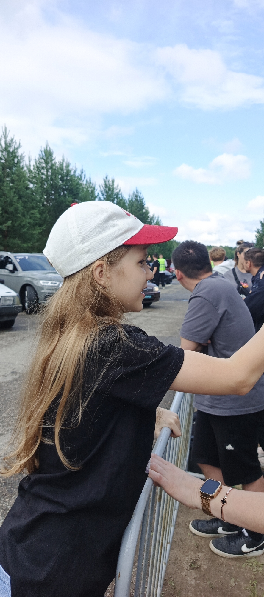 A young girl with long blonde hair and a white and red baseball cap is leaning on a metal fence, looking to her right. She is wearing a black t-shirt. In the background, several people are visible, as well as cars and a line of trees under a cloudy sky. The scene appears to be outdoors, possibly at an event or gathering. The location in the city of Chaikovsky, Russia is indicated by the presence of a blue and white license plate on a dark-colored car in the background.