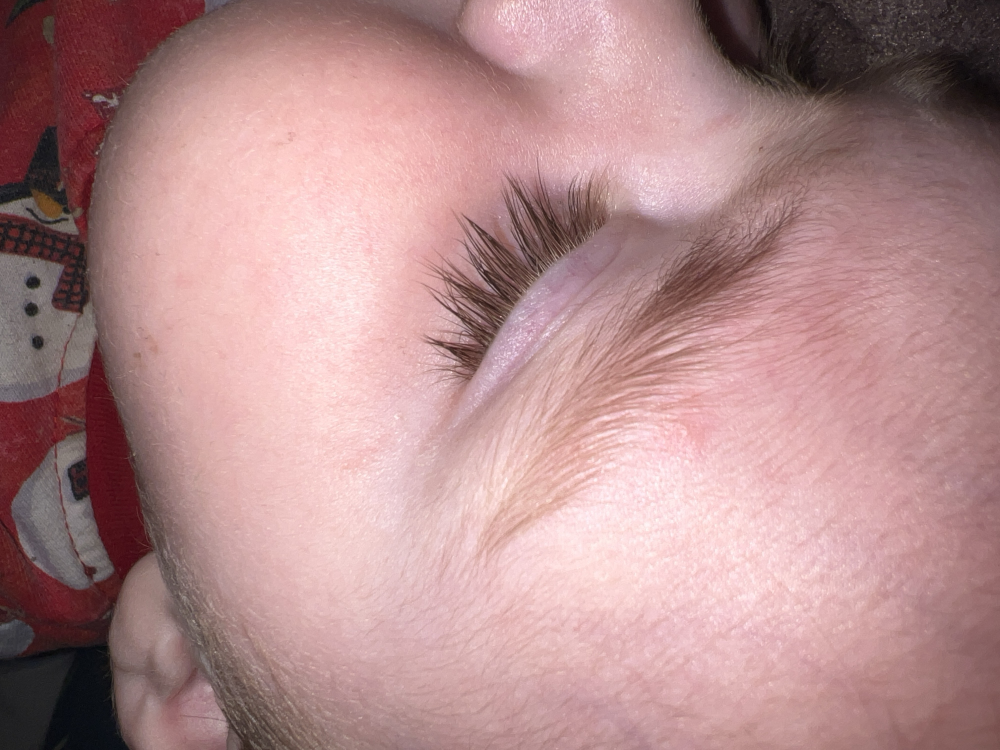 A close-up image shows a child's face. The child is sleeping, with their eyes closed and long eyelashes visible. A red garment with a snowman design is partially visible in the upper left corner. Fine hair is visible on the child's forehead and cheek. The skin is pinkish in tone.