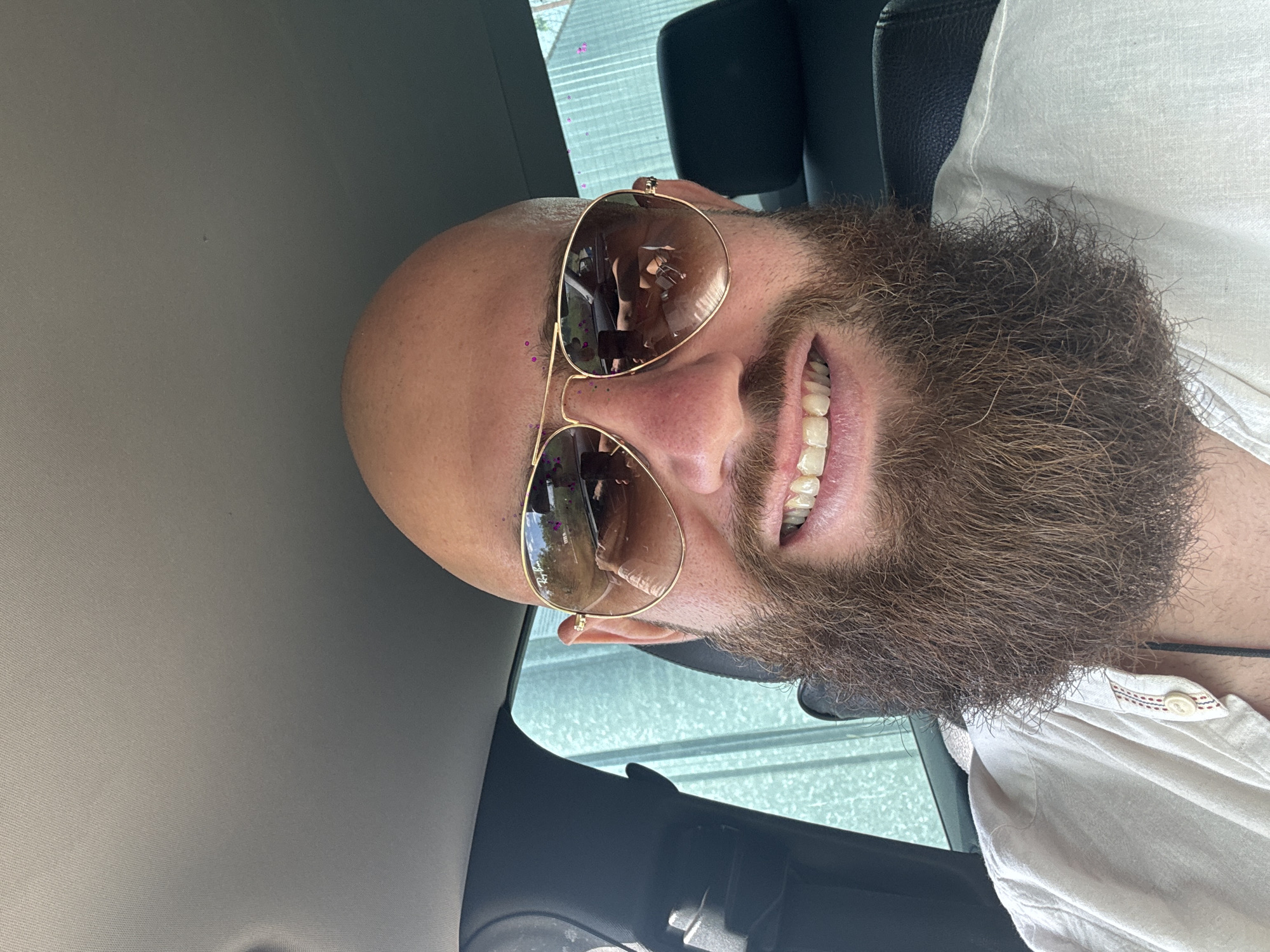 A man with a beard and sunglasses is sitting in a car. He is wearing a white shirt. The background shows a glimpse of a city street with buildings. The location is Unknown, Unknown.