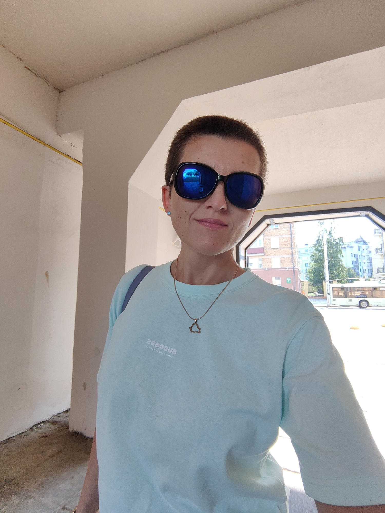 A woman with short brown hair and a light blue t-shirt stands indoors, looking at the camera. She is wearing large, dark sunglasses with blue reflective lenses and a delicate gold necklace with a triangular pendant. Her t-shirt has a white graphic print on the left side that reads "SUCCESS". A blue strap from a bag is visible over her left shoulder. The background shows an opening to the outdoors, where a street scene with buildings, trees, and a trolleybus can be seen. The woman appears to be in a covered walkway or parking area with white walls and a concrete floor. The lighting suggests it is daytime. Based on the presence of a trolleybus which is common in Minsk, Belarus, and the architectural style of the buildings in the background, this scene is likely located in the city of Minsk, Belarus.