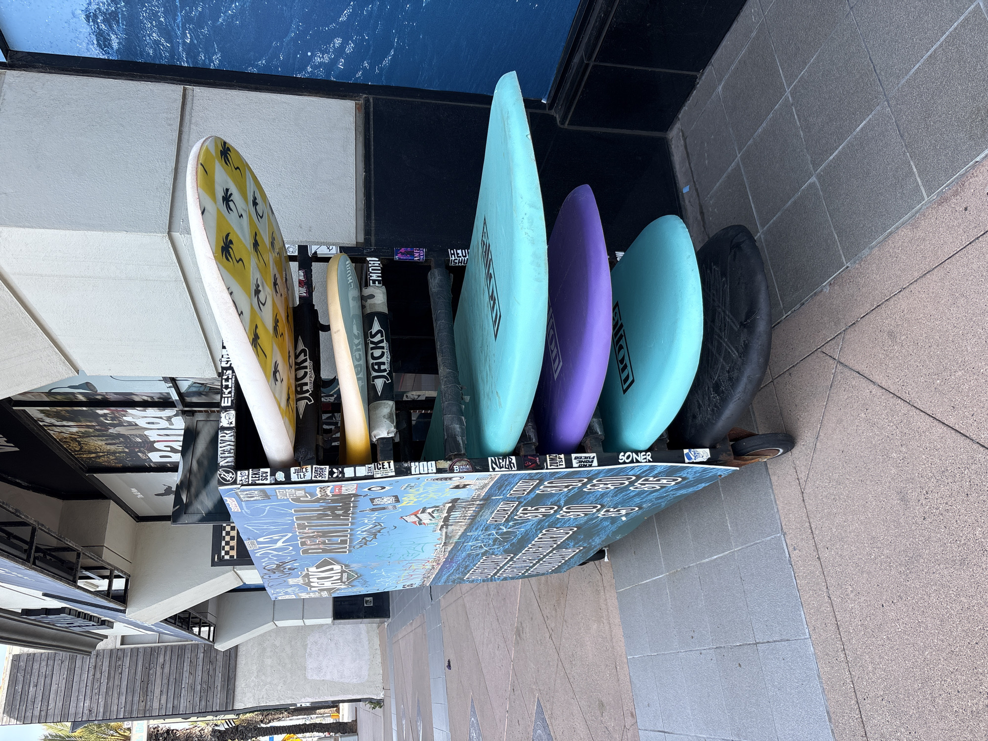 This image, rotated, displays a colorful array of surfboards vertically held in an outdoor rack, likely associated with a surf shop or rental in Huntington Beach. Six surfboards are visible. On the far left, a white surfboard features a distinctive yellow diamond pattern, each diamond containing a black palm tree silhouette. Next to it is a light, possibly beige, surfboard. These are followed by three vibrant boards: a light turquoise, a purple, and another light turquoise, all of which display "ALLON" printed vertically. The final surfboard on the right is dark grey or black, possibly covered.
The metal surfboard rack is extensively covered in an assortment of stickers and graffiti. Visible text from these decals includes "JACKS SURFBOARDS" (appearing multiple times), "NEWREKIS", "EMON", "HEU ISHU", "SONER", "SCV", "TLF", "3XK", "BOR", "CHUR", "ANE", "LEKO", "BA", "NEZE", and "SIDH." The lower front panel of the rack is a large blue board depicting a stylized ocean scene with waves, over which white text and additional graffiti are layered, making some parts illegible, though "REMIND" can be partially seen.
In the background, a building facade with windows and dark framing is visible. Behind the surfboards, there's a dark blue wall or display panel featuring a textured design resembling ocean water. The ground is paved with light-colored square tiles. The scene is brightly illuminated, indicating daytime conditions. The surfboards and the overall aesthetic strongly suggest a coastal, surf-centric environment.