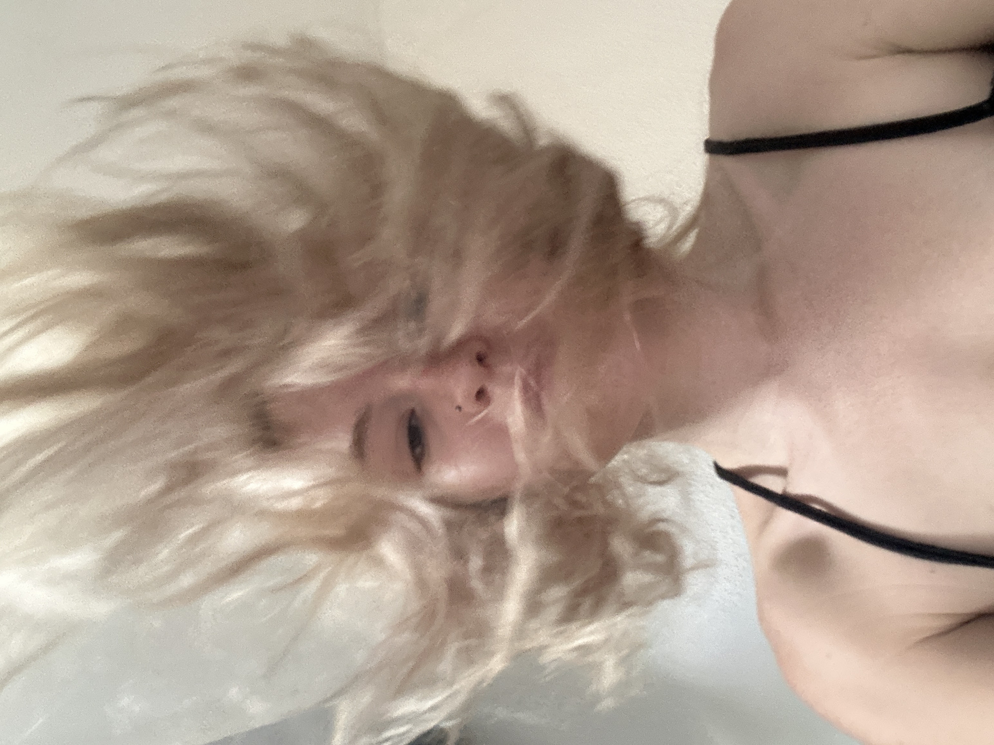 The image shows a close-up, slightly blurry, upward-angle shot of a young person with blonde hair that is in motion, obscuring much of their face. The person appears to be wearing a black camisole or bra. The background is a plain, light-colored wall. The image does not contain any discernible elements that would indicate a specific location within Tbilisi, Georgia.