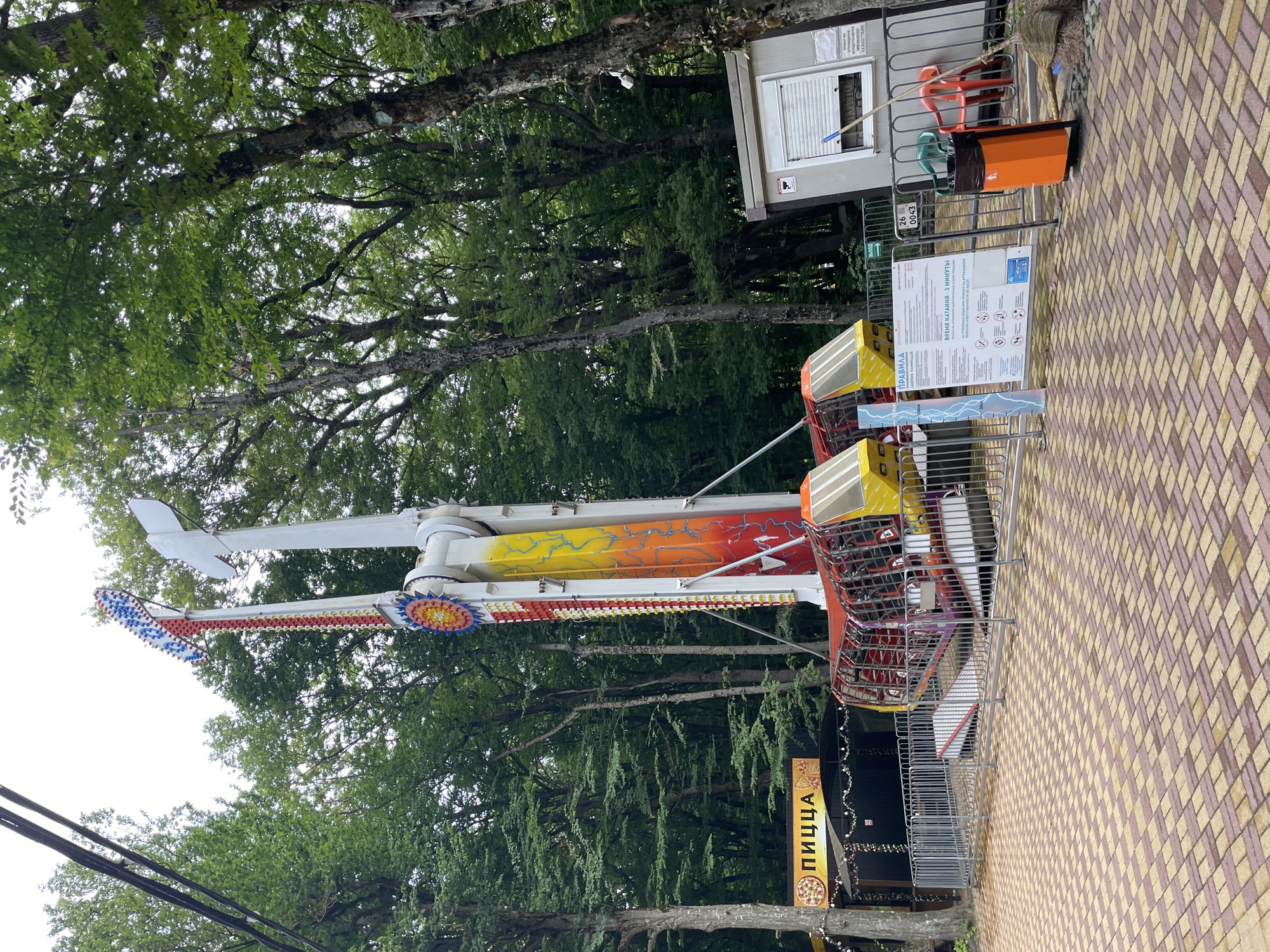 The image shows an amusement park ride in an outdoor setting, likely during daylight hours. The ride is a tall, vertically swinging pendulum-style attraction with two passenger pods, each seating several people. The ride structure is predominantly white, with a colorful section featuring red, yellow, and orange flames and a circular emblem with blue and yellow accents. In the foreground, a patterned paved area transitions to a metal safety fence. Behind the fence, a small building with a window and a sign with Russian text is visible. To the left of the ride, a banner with the word "ПИЦЦА" (Pizza) in Russian indicates a food vendor. Tall, lush green trees surround the amusement park area. The sky appears overcast. No people are visible in the image. The overall scene depicts an operational amusement park attraction in a natural, tree-lined environment.