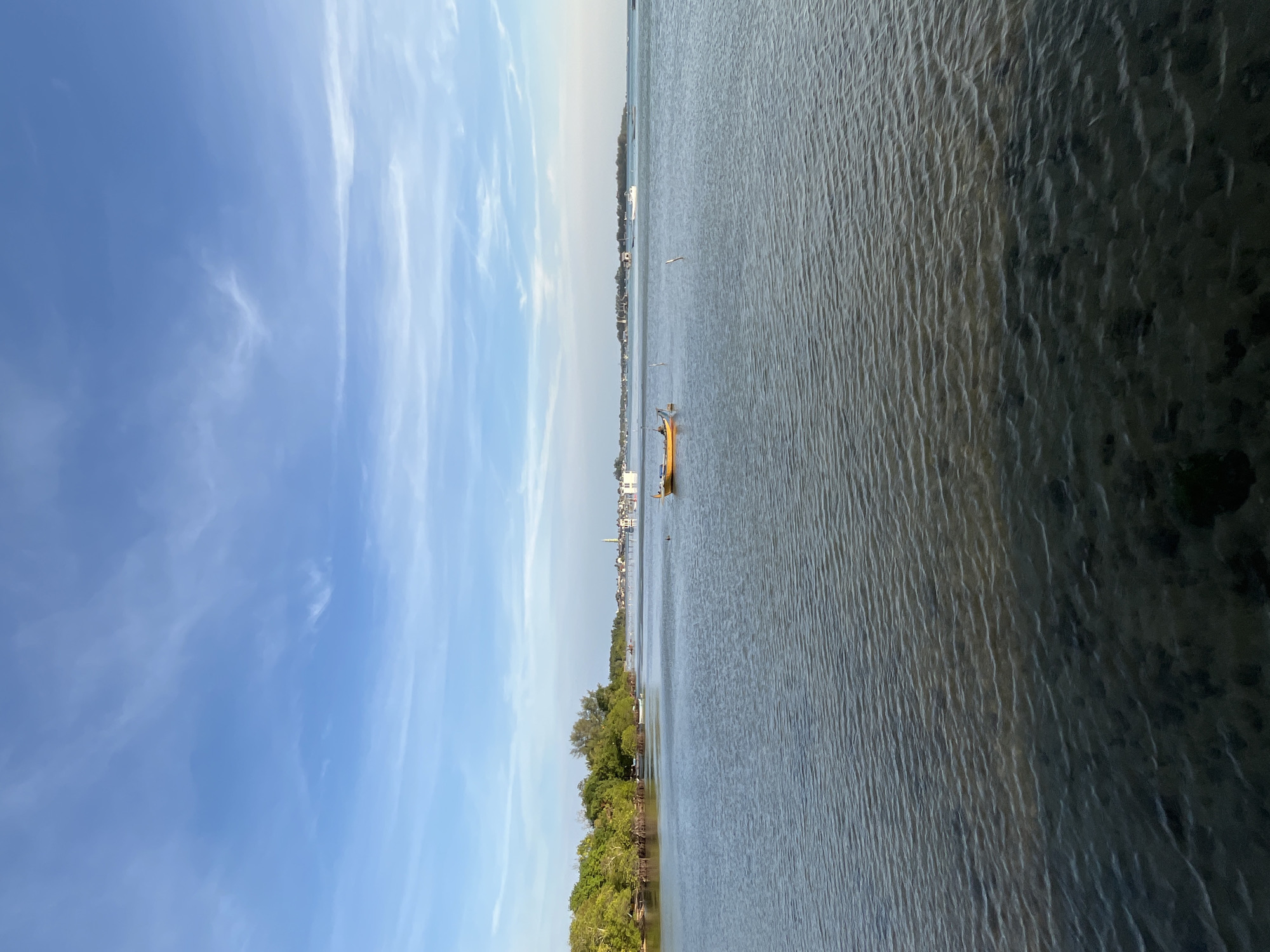 This is a photograph taken from a shoreline in the city of Karimunjawa, Indonesia, looking out over a calm body of water. The sky is a bright blue with scattered wispy white clouds. The water is relatively still, with gentle ripples on the surface, and appears shallow near the shore, revealing a dark, silty bottom.
On the left side of the frame, a lush green coastline is visible, densely populated with trees. In the distance, along the horizon, a cluster of buildings and structures can be seen, suggesting a settlement or town. Several boats are present on the water. The most prominent is a small, yellow, motorized fishing boat situated slightly right of the center of the frame, appearing to be stationary. Another smaller, darker boat is visible further in the distance, closer to the shoreline. A thin, vertical pole or marker stands in the water between the two boats. The overall scene depicts a tranquil coastal environment.