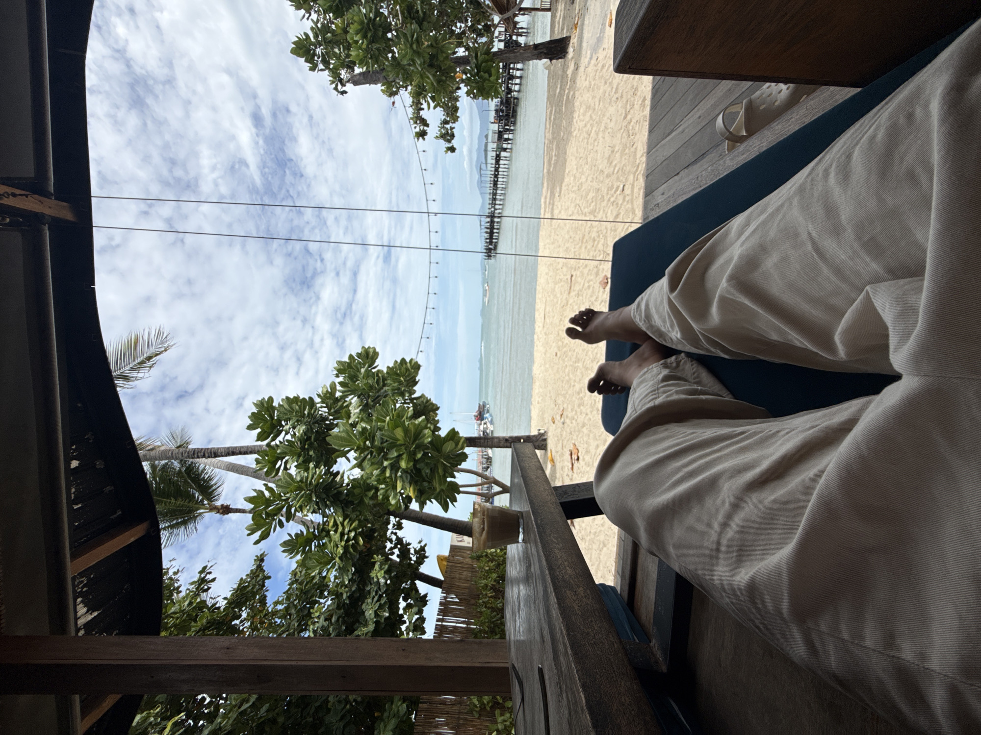 The image shows a first-person perspective of someone relaxing on a lounge chair on a sandy beach. The person is wearing light-colored pants, and their bare feet are visible. In the foreground, there is a wooden table with a glass of beverage on it. Beyond the table, the beach stretches to the ocean, where a long pier extends into the water. Several boats are visible in the distance, and palm trees and lush green foliage frame the scene. The sky is partly cloudy, with a bright, hazy sun. The setting appears to be a beachfront cafe or resort in the Ko Samui District of Thailand.