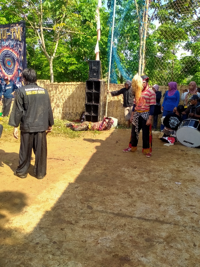 An outdoor scene in Plosorejo, Indonesia, depicts a cultural performance or event on a dirt ground under bright daylight. In the foreground, a male figure stands with his back to the camera, wearing a black uniform consisting of a long-sleeved top with white text "kesenian reog SIMO MUDHO MANGGOLO" on the back and black wide-legged pants. To his right, another male performer stands facing slightly left, adorned in a vibrant costume featuring a large white/blonde-colored fibrous headpiece, a red and white striped t-shirt, black pants with red and yellow accents, and a black and white checkered sash around his waist, complemented by red sneakers.
In the mid-ground, slightly to the left, an individual in traditional attire is lying prone on the grassy patch at the base of a stacked sound system. This system consists of two large black speaker cabinets. Immediately behind the speakers and the lying figure, a woven bamboo fence serves as a backdrop, partially obscured by green foliage and trees in the background. Blue netting is visible behind the fence and trees on the right side.
To the right of the speaker system, a male individual in dark clothing stands with his left arm extended outwards. Further to the right and in the background, a small crowd of spectators observes the scene. This group includes several women wearing various colored headscarves (hijabs) and men, some of whom are seated. A large white bass drum with the word "TRITON" printed on its head is visible on the far right, near the seated individuals. A distinct shadow line casts across the dirt ground in the foreground, indicating the position of the sun. A banner with text "TW-TM" and "RANTING" along with a circular emblem is partially visible on the far left, behind the first performer.